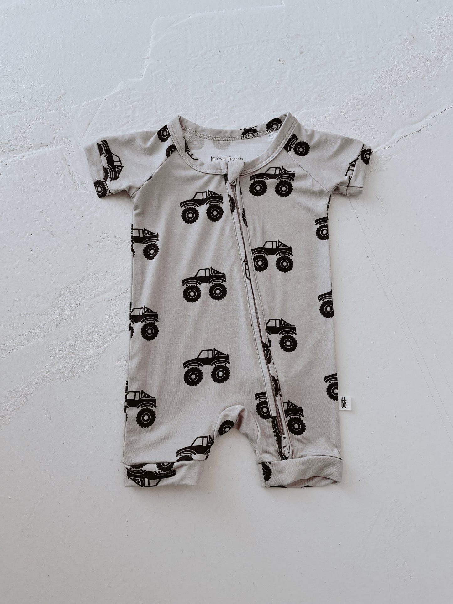 The forever french baby Shortie Bamboo Zip | Monster Truck is a short-sleeved, light gray romper with breathable fabric and black monster trucks, finished with a zipper—perfect for little ones needing comfort and style.