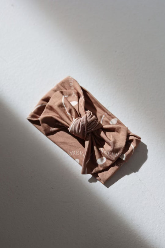 The Bamboo Head Wrap | ff Desert Clay Smile by forever french baby, a folded light brown fabric headband with white polka dots and a center knot, lies on a gray surface illuminated by soft, natural light.