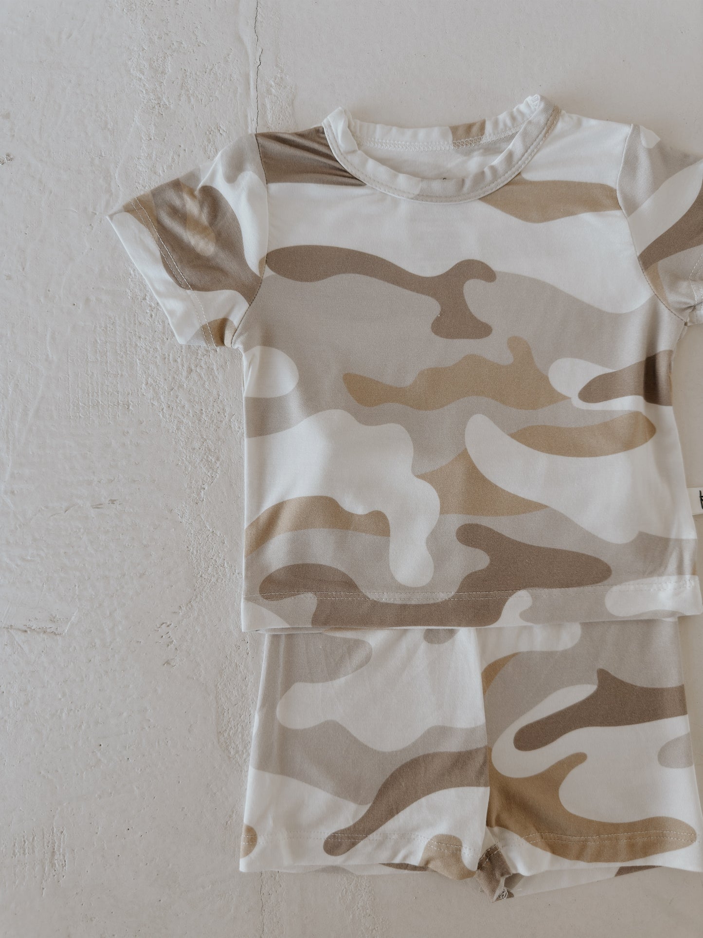 The Bamboo Short Set | Camo 2.0 by forever french baby features a beige, tan, and brown camo print in breathable fabric. This comfy short-sleeve set is shown laid flat on a textured light surface.