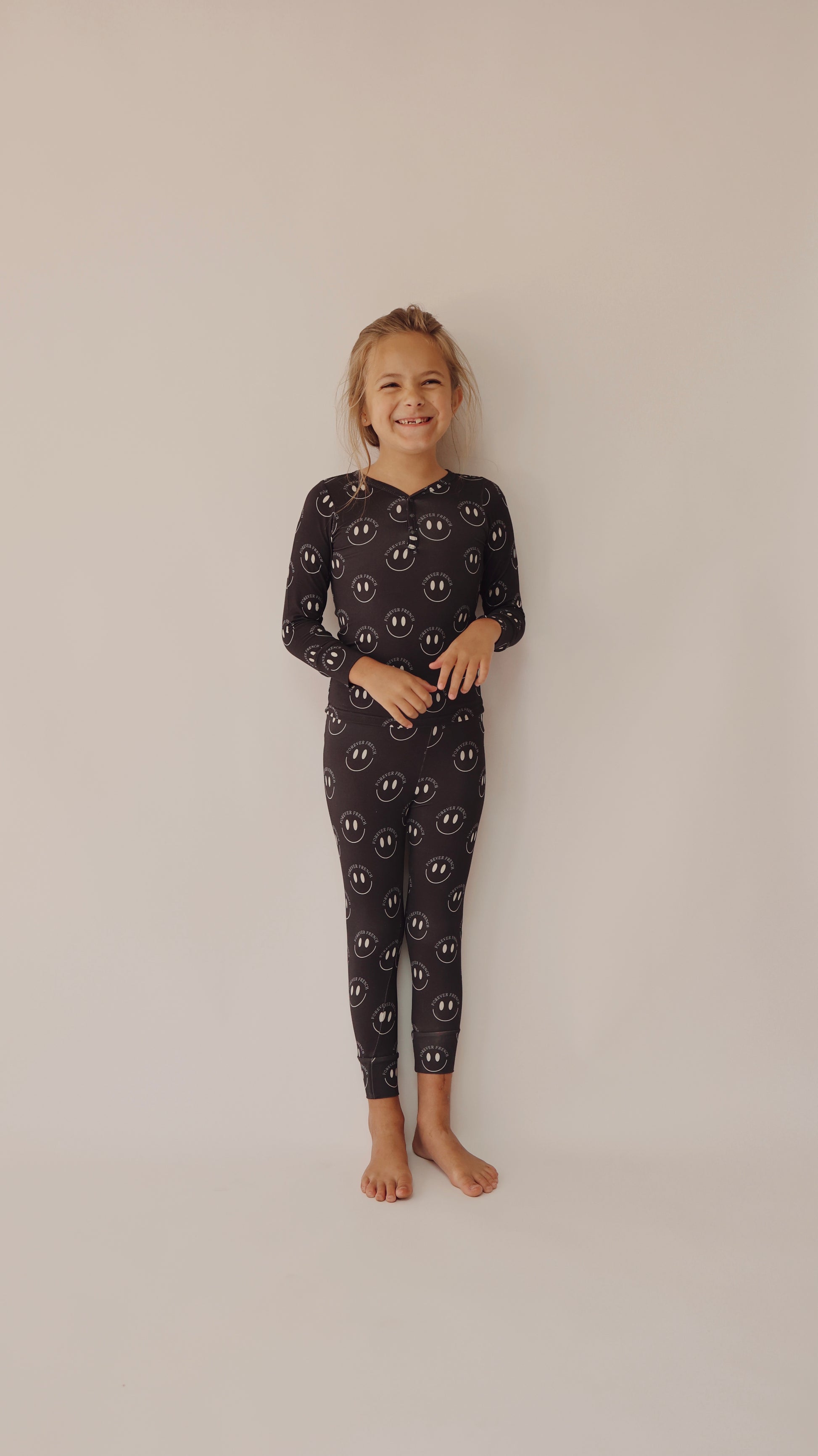A young child stands barefoot against a plain background, smiling widely. They are wearing the Bamboo Two Piece Pajamas in Charcoal & White from forever french baby, covered in white smiley face patterns. The child has light-colored, tousled hair and a relaxed, happy expression on their face.