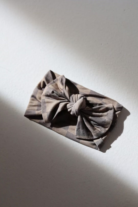 The "Bamboo Head Wrap | Faded Brown Checker" by forever french baby, featuring a knotted bow design and a brown and beige checkered pattern, is displayed on a light gray surface with soft natural light casting shadows. Made from soft bamboo material, this adjustable head wrap is perfect for babies and toddlers.