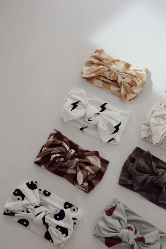 A beautifully arranged collection of eight adjustable head wraps from Forever French Baby, perfect for babies and toddlers, displayed on a light surface. Each head wrap in this set, called Bamboo Head Wrap | ff Desert Clay Smile, is crafted from soft bamboo material and showcases unique patterns like plaid, lightning bolts, yin-yang symbols, and abstract designs in neutral earthy tones such as browns, whites, and grays.