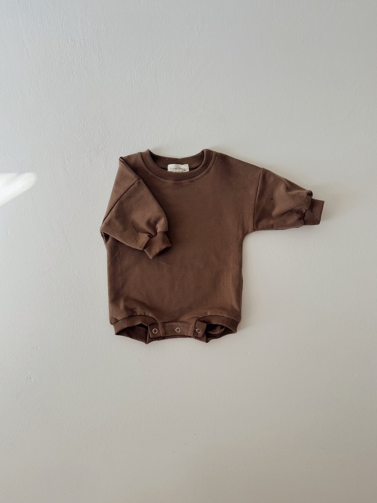 The Sweatshirt Romper | Hot Cocoa by forever french baby, made from brown organic cotton with long sleeves and bottom snaps, is laid flat against a light gray wall.