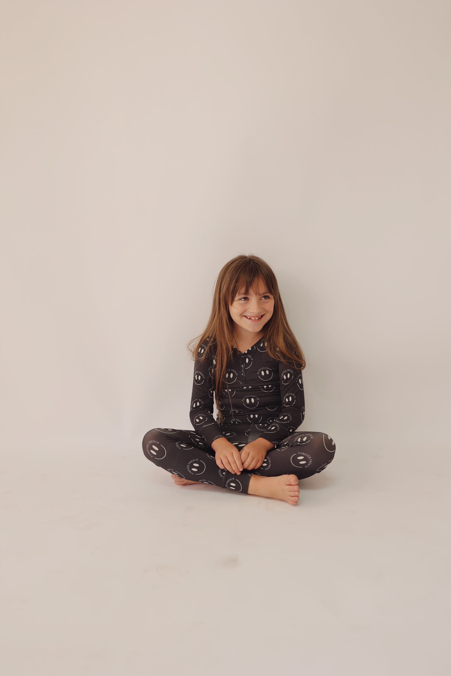 A young girl with long brown hair sits cross-legged on the floor against a plain white background. She is wearing the Bamboo Two Piece Pajamas in Charcoal & White ff Smile by forever french baby. Her breathable sleepwear, adorned with white smiley face patterns, allows her to smile comfortably and look off to the side.