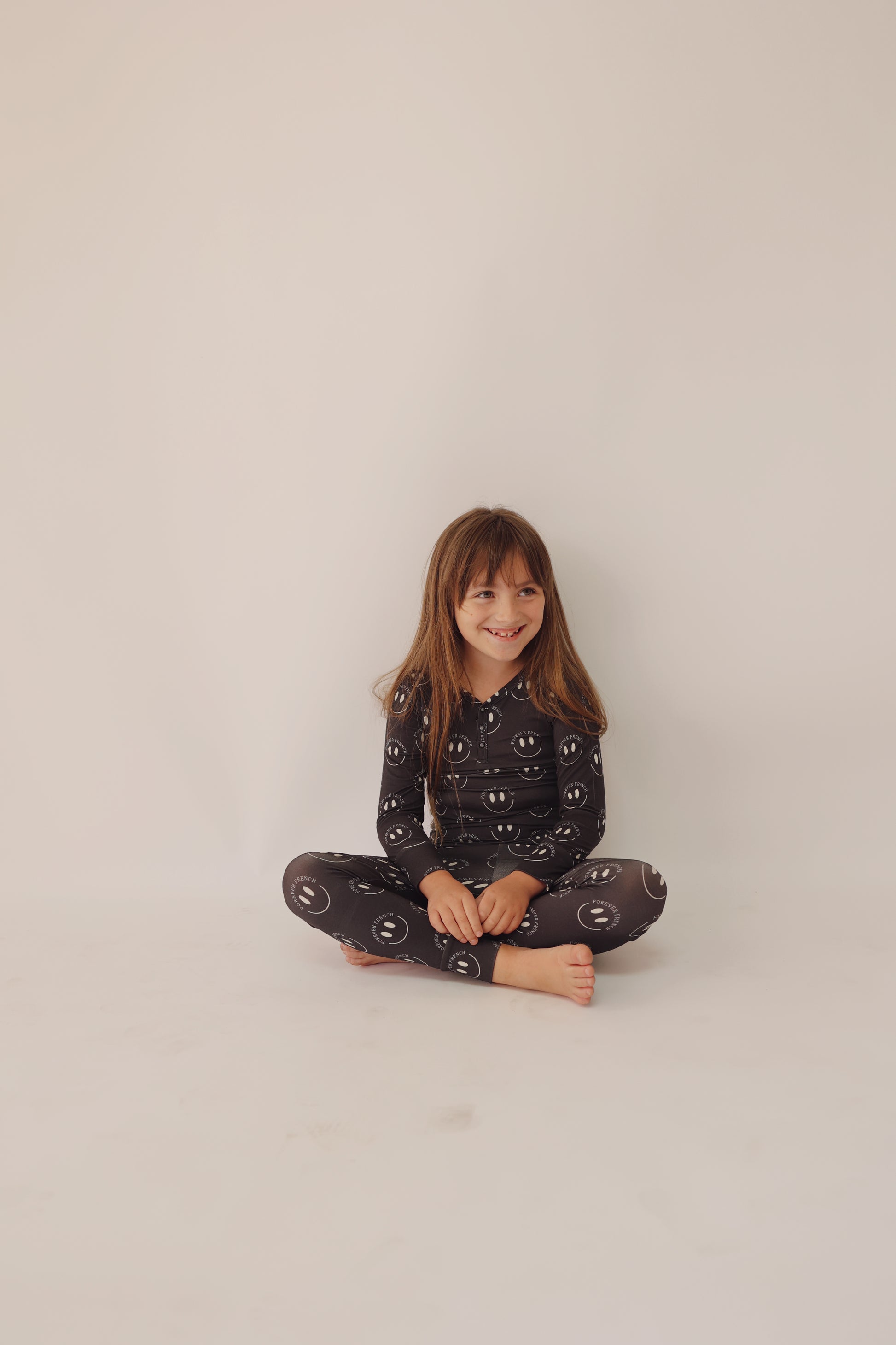 A young girl with long brown hair sits cross-legged on the floor against a plain white background. She is wearing the Bamboo Two Piece Pajamas in Charcoal & White ff Smile by forever french baby. Her breathable sleepwear, adorned with white smiley face patterns, allows her to smile comfortably and look off to the side.