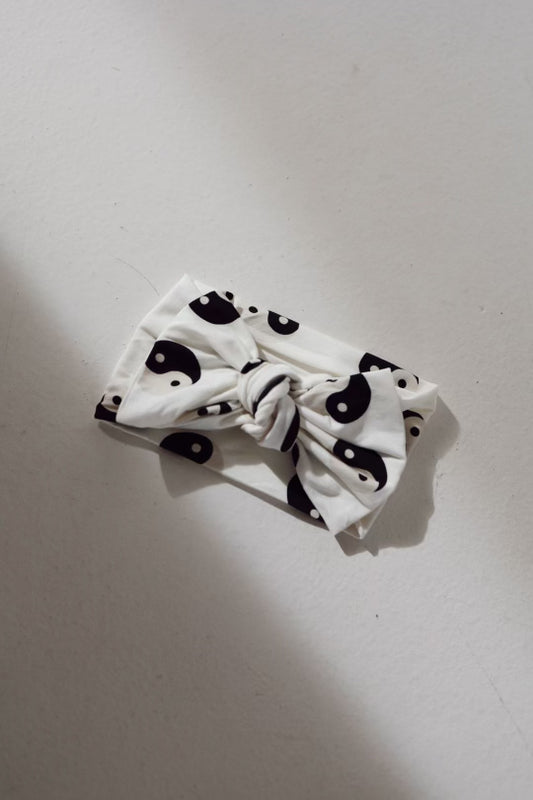 A Forever French Baby Bamboo Head Wrap in black and white yin-yang pattern, featuring a knotted bow in the center, is placed on a smooth, light-colored surface. This elegant piece seamlessly integrates charm and style.
