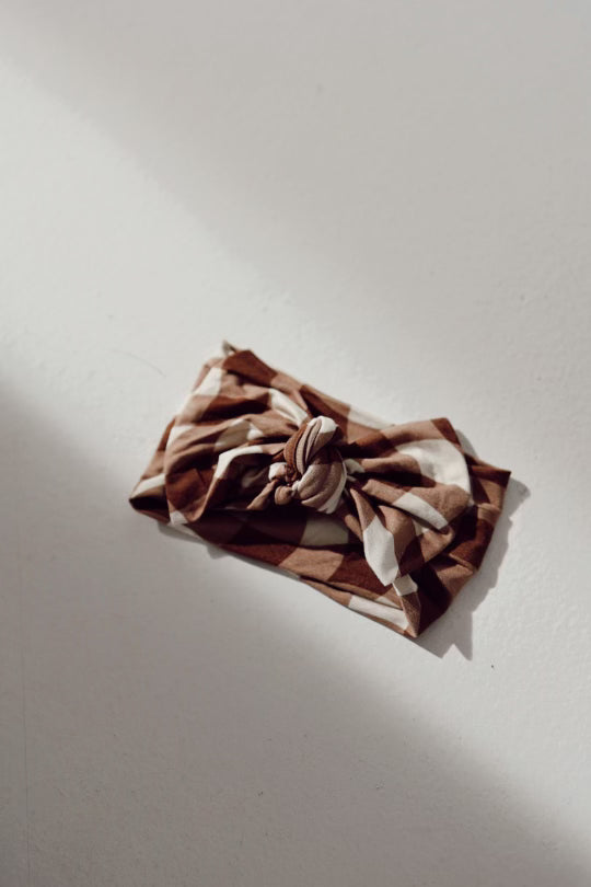 A Bamboo Head Wrap | Groovy Gingham by forever french baby, featuring a large knotted bow in the center and a charming brown and white checkered pattern, rests on a light grey surface. The sunlight cast beside it creates a soft shadow, enhancing the texture's depth.