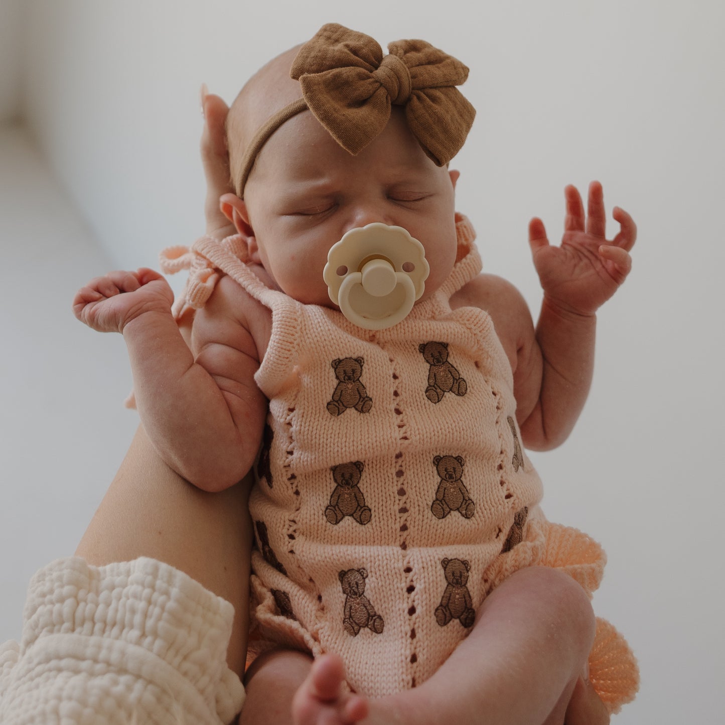A newborn baby girl sleeps peacefully with a pacifier, cradled in an adult’s arm, wearing forever french baby’s Knit Ruffle Romper | Lottie and a brown bow headband.