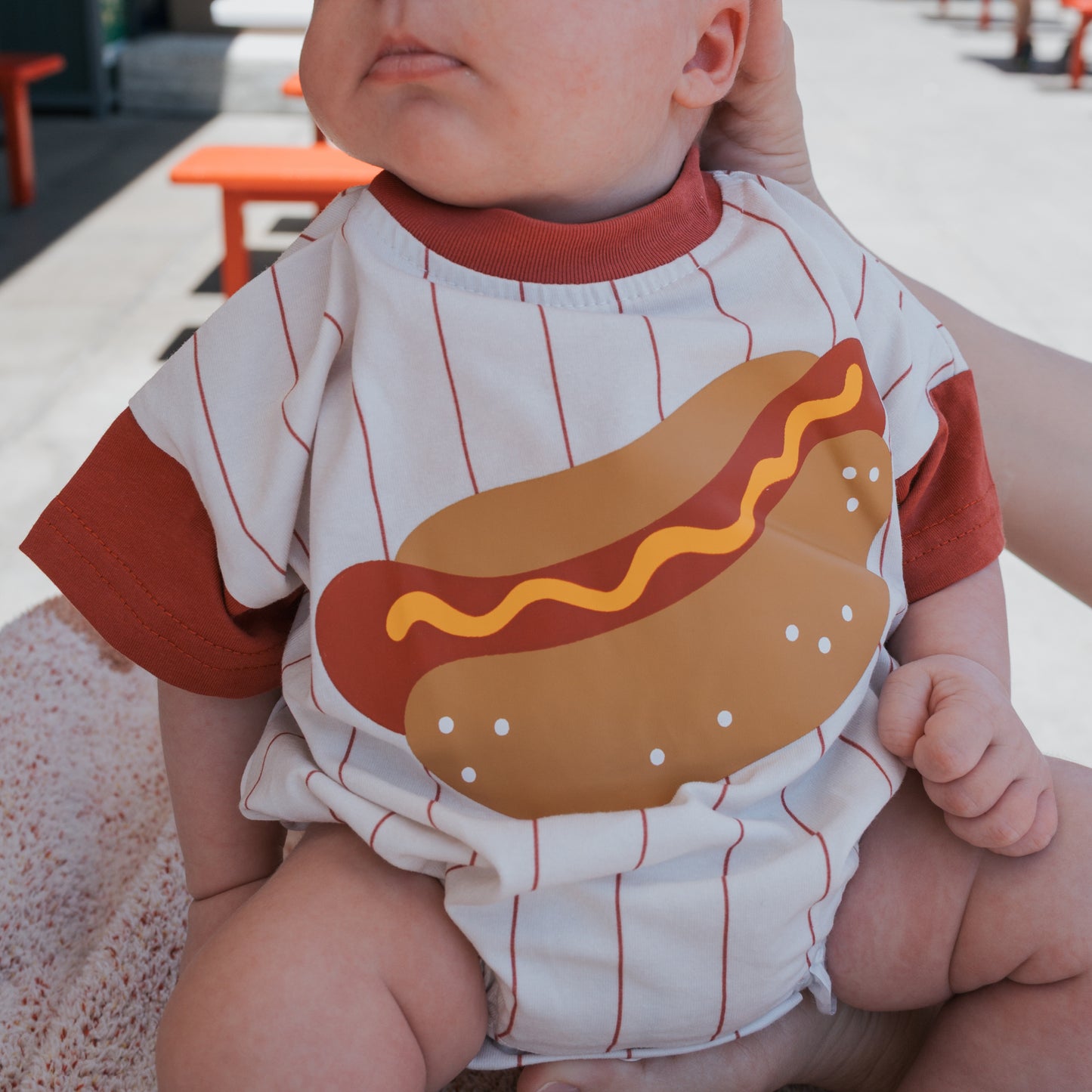 A baby wears the forever french baby Short Sleeve Romper | Hot Dog, a cute white and red cotton romper with a big hot dog graphic—bun, sausage, mustard—perfect for outdoor fun while being held by an adult.