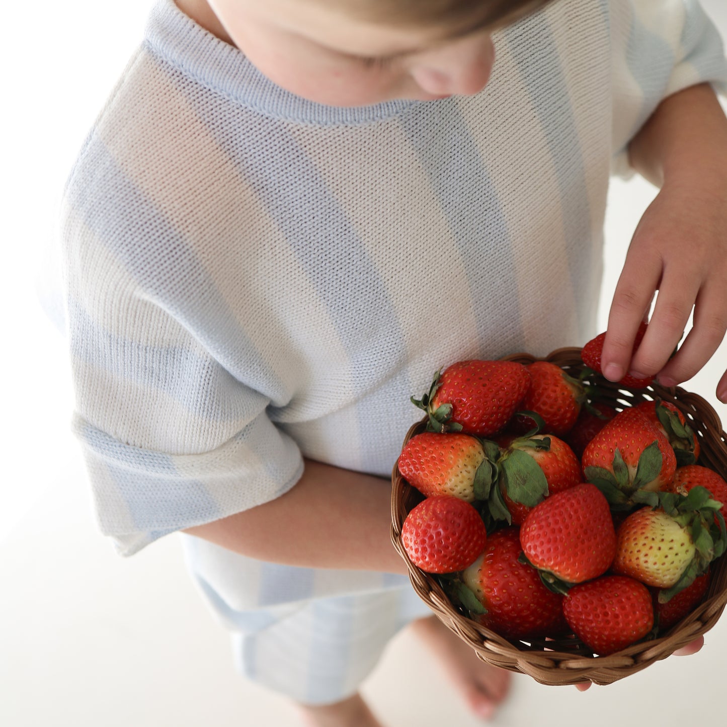 A child in a light blue and white Knit Short Set | Benny by forever french baby holds a basket of fresh strawberries, reaching in to pick one. The background is bright and minimal.