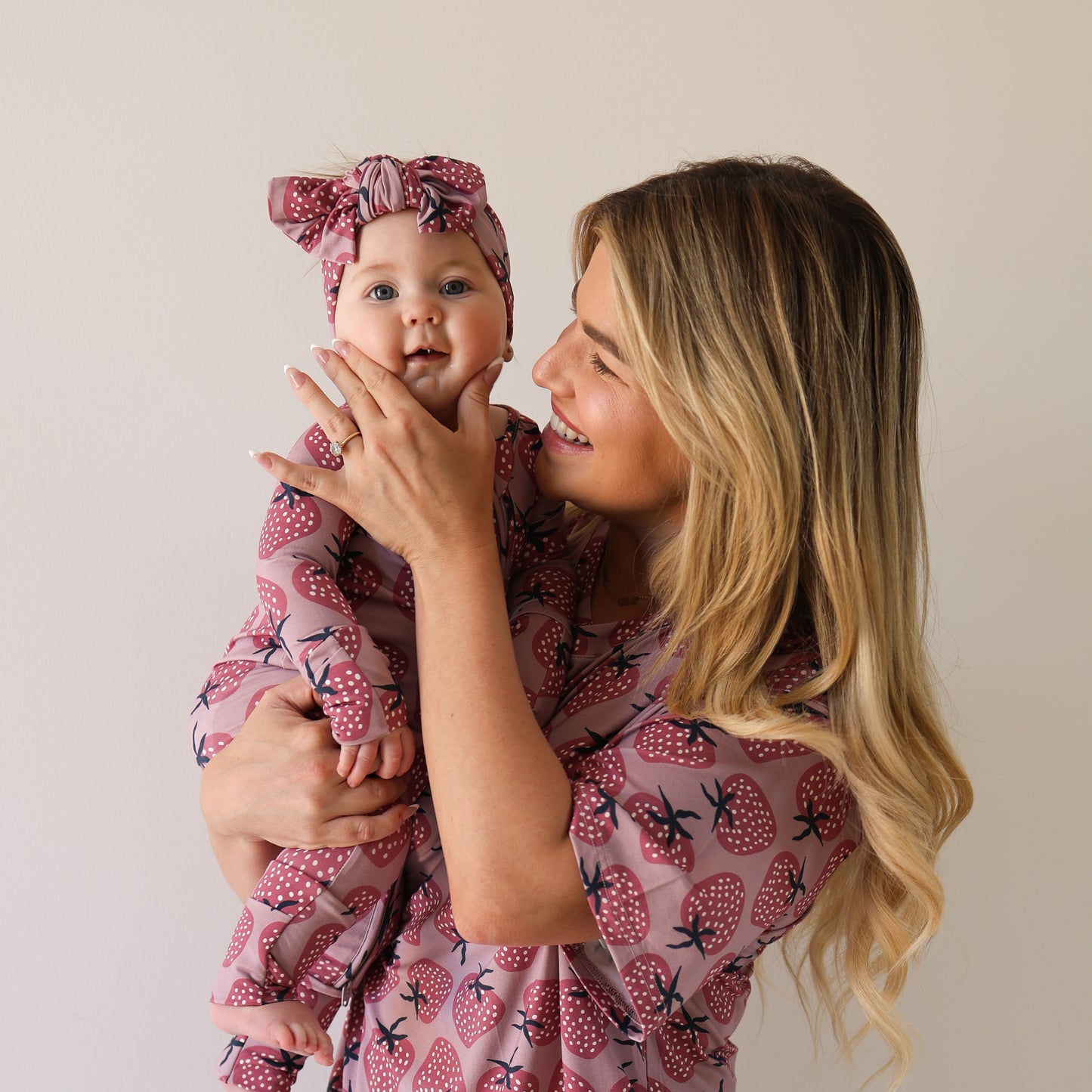A smiling woman with long blonde hair holds a baby as they wear matching forever french baby Bamboo Zip Pajamas | Matilda in a strawberry print. The baby, wearing a bow headband, looks at the camera while the woman gazes at her affectionately.