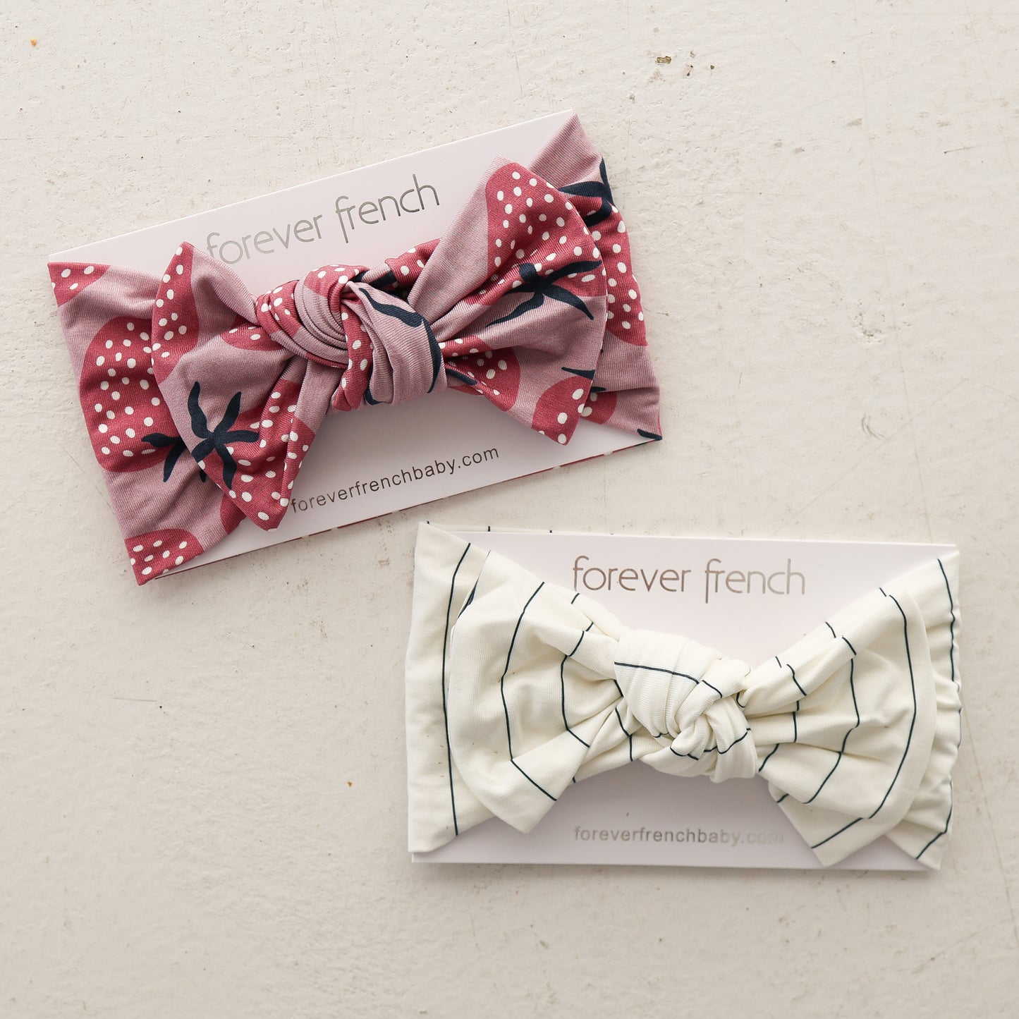 Two Bamboo Head Wrap | Matilda headbands by forever french baby are shown on branded cards: one is pink with white polka dots and florals, the other cream with thin black stripes, all on a light, textured surface.