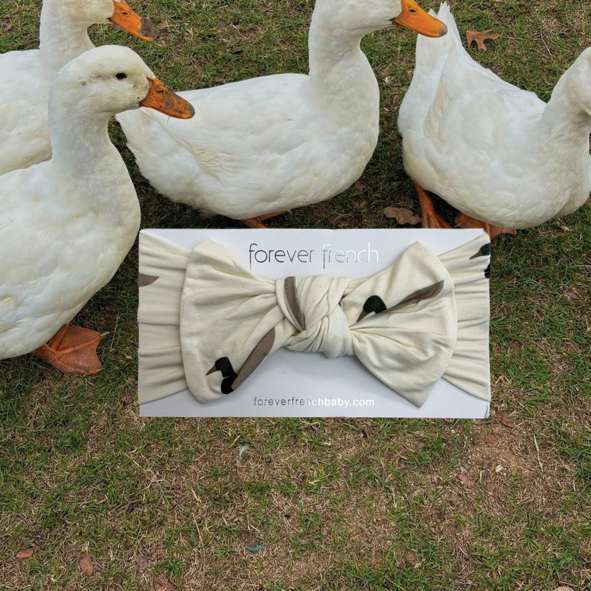 Four white ducks stand on grass behind a forever french baby Bamboo Head Wrap | Mallard, adorned with a bow and placed on the ground in front of them. This charming head wrap showcases a stylish mallard-inspired white and black pattern.