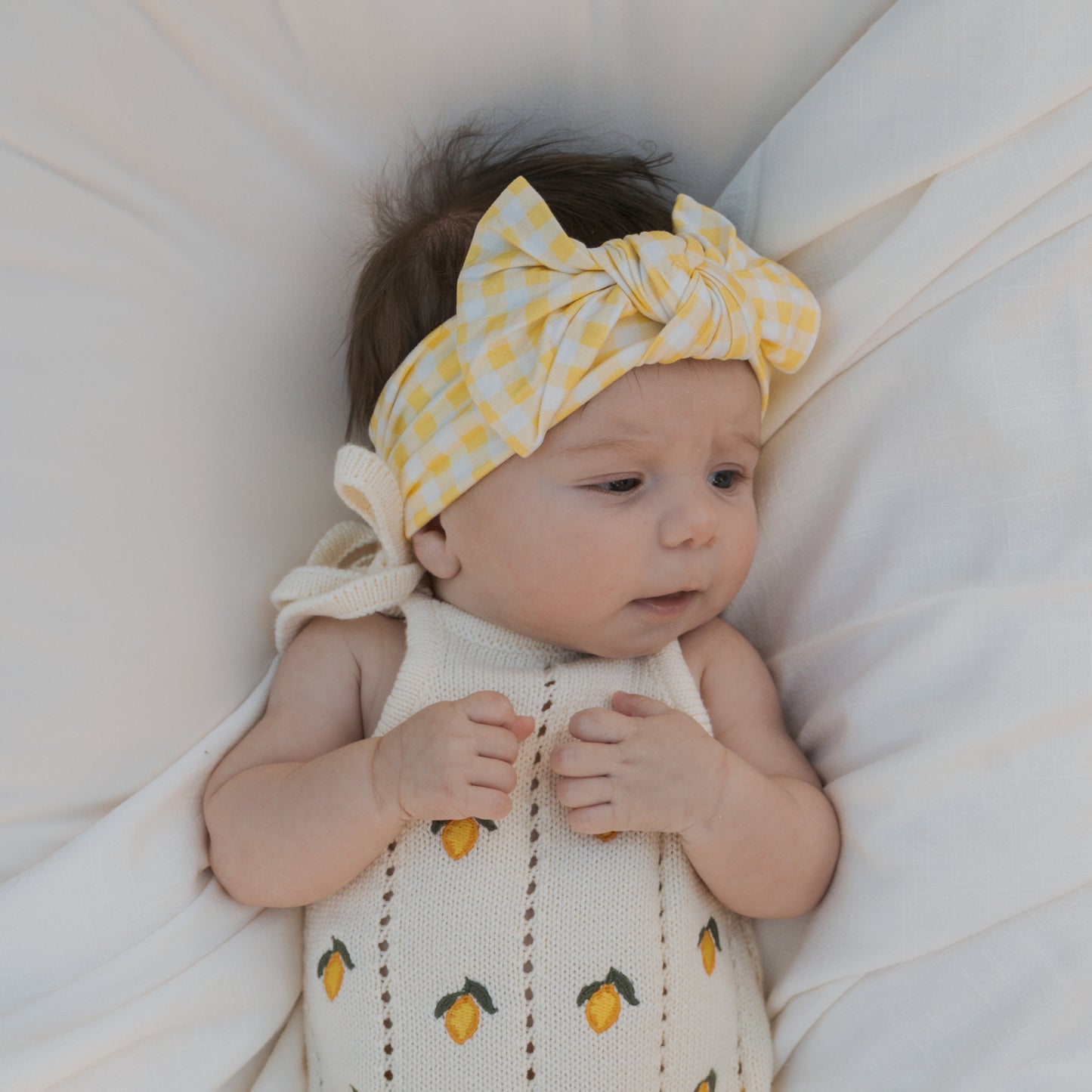 A baby lies on a white blanket, wearing a cream knit outfit with small lemons and a forever french baby Bamboo Head Wrap | Citron featuring a large bow, looking to the side with hands resting on their chest.