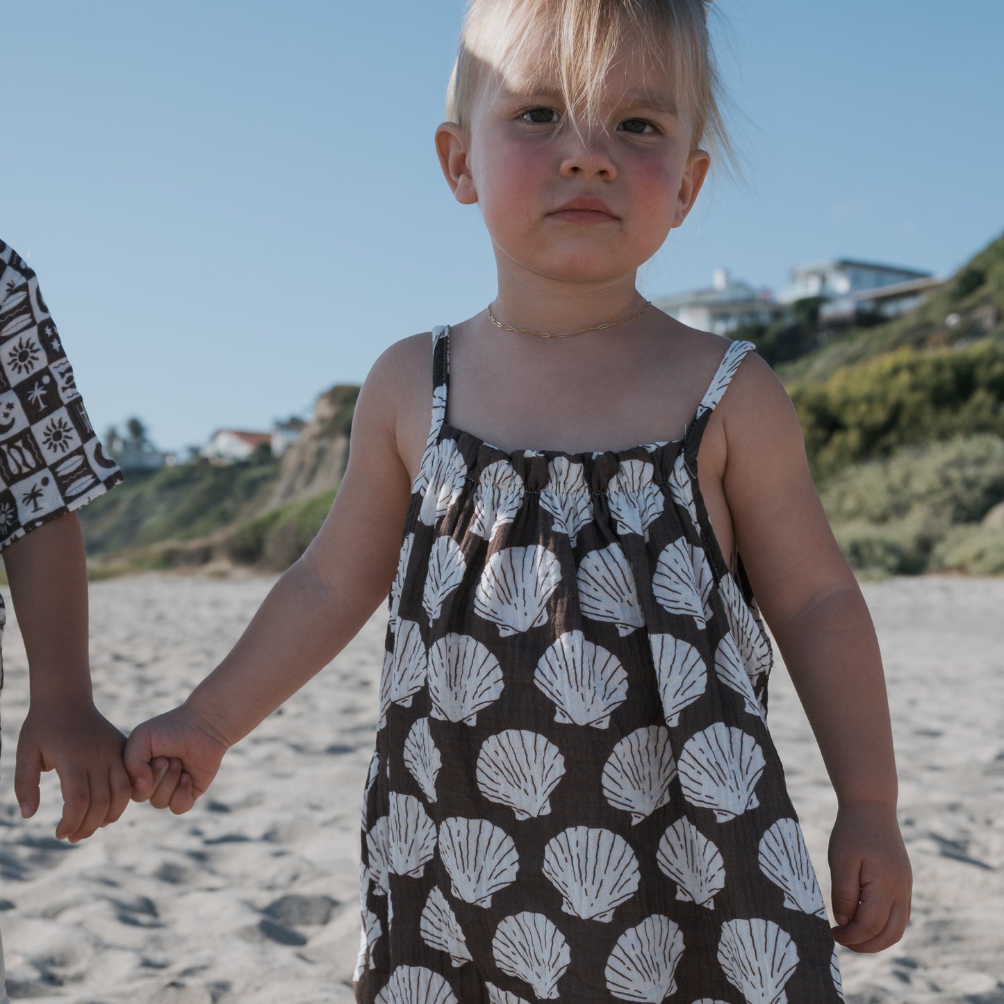 A young child in the forever french baby Child Muslin Sleeveless Dress | Seashells stands on a sandy beach, holding another person’s hand. In the background are hillside houses and greenery under a clear sky.