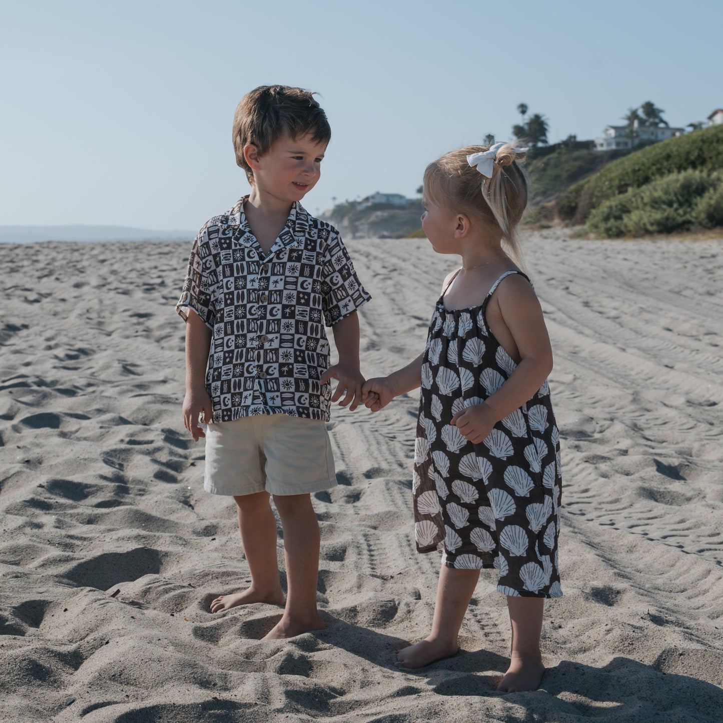 A young boy and girl smile at each other, holding hands on a sunny beach. The girl wears the forever french baby Child Muslin Sleeveless Dress | Seashells, with houses and greenery in the background.