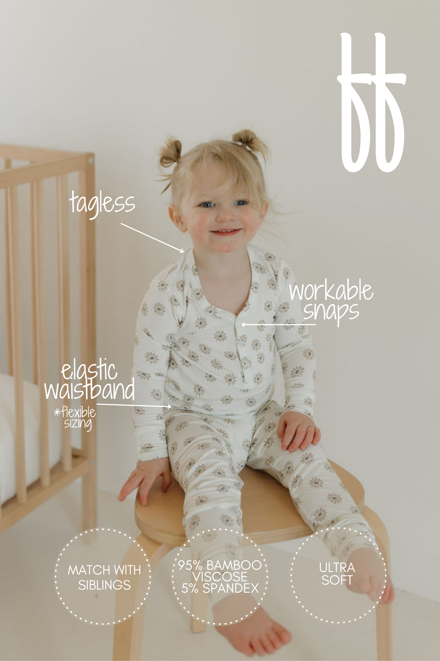 A toddler with pigtails sits on a wooden stool, wearing forever french baby's "Bamboo Two Piece Pajamas | Daisy Fields" featuring a floral-patterned hypo-allergenic design. Highlights include tagless feature, workable snaps, elastic waistband, and breathable fabric. A crib is partially visible in the background.