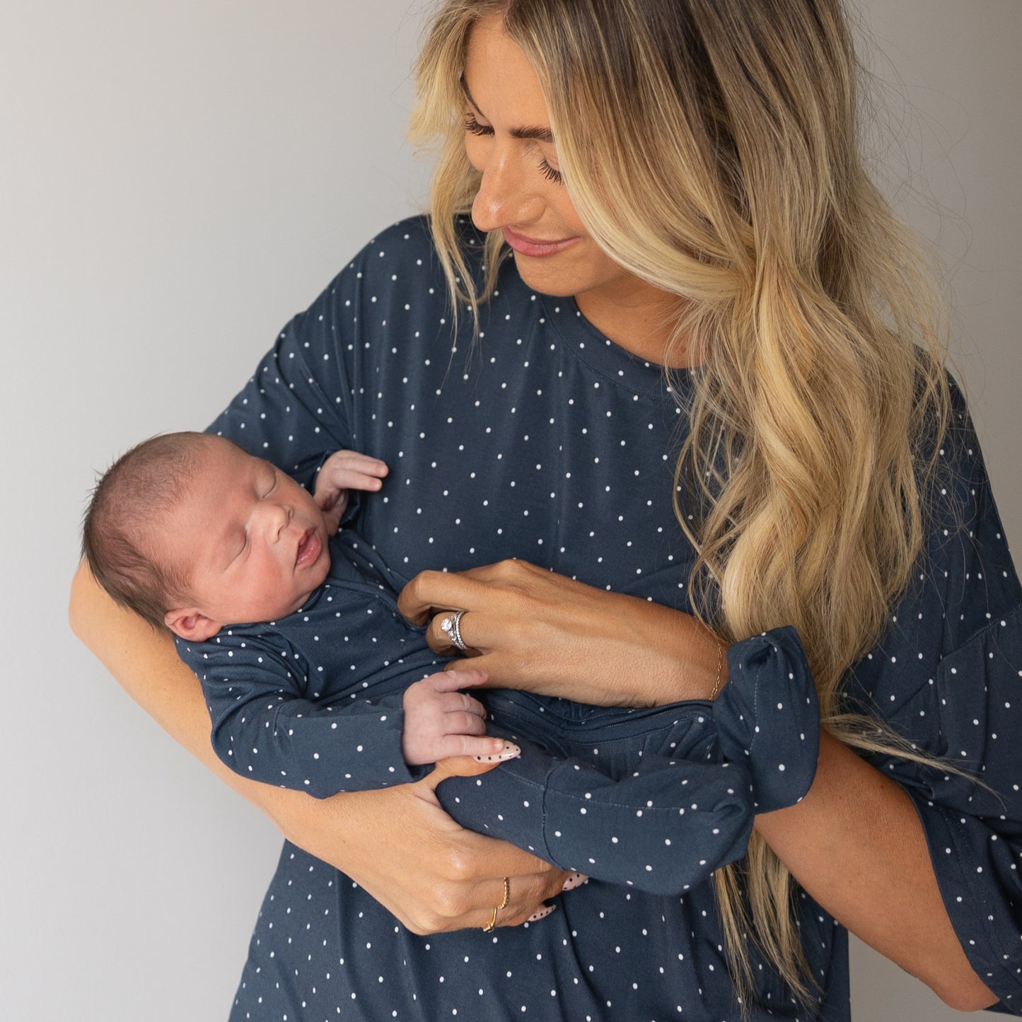 A woman with long blonde hair holds a sleeping newborn, both in matching forever french baby Bamboo Zip Pajamas | Emery in navy blue with white polka dots. She looks down and smiles gently, enjoying their cozy bamboo sleepwear.