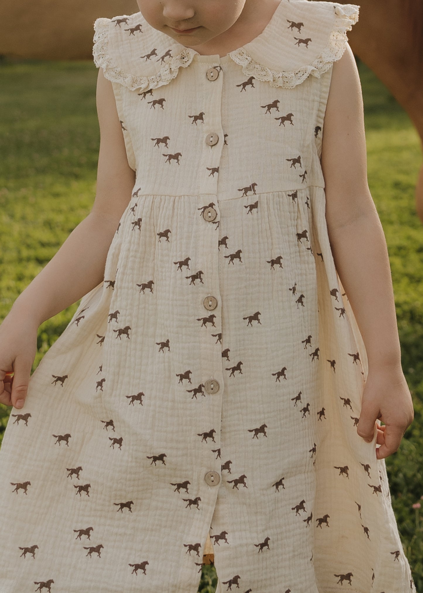 A child in the forever french baby Child Muslin Dress | Wild Horses stands on green grass, looking down with arms slightly out, wearing a sleeveless cream dress with lace collar and brown horse prints.