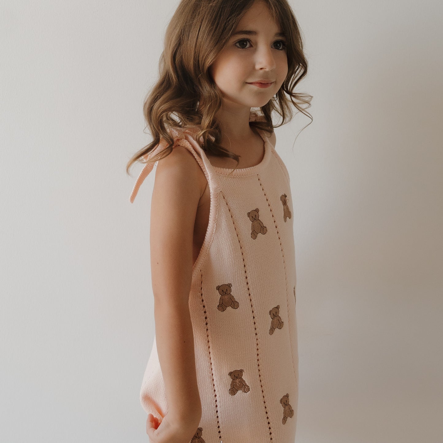 A young girl with wavy brown hair wears the forever french baby Knit Dress | Lottie— a sleeveless 100% cotton pink knit dress adorned with embroidered teddy bears, standing against a plain white background.