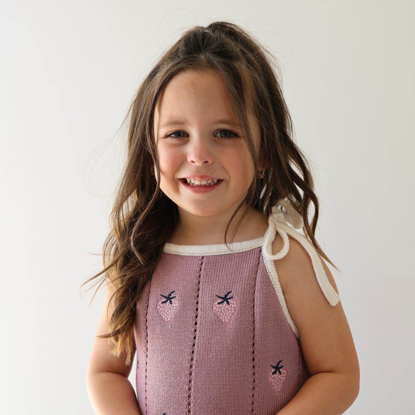 A young girl with long brown hair wears the forever french baby “Knit Dress | Matilda,” a purple sleeveless dress with adjustable tie straps, and smiles at the camera against a plain white background.