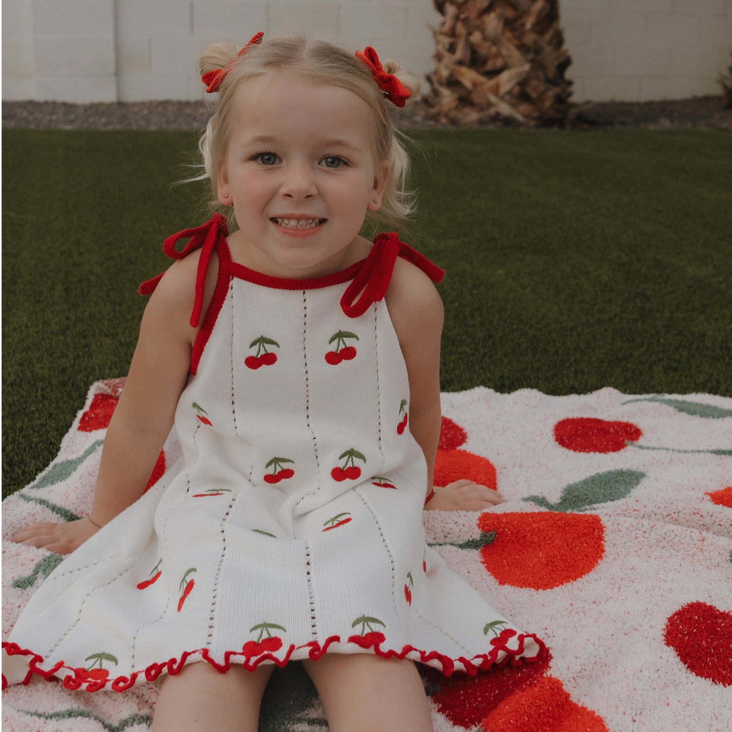 A young blonde girl smiles at the camera outdoors, sitting on a cherry-print blanket in the forever french baby Knit Dress | Cherry—white 100% cotton with red cherry designs, adjustable tie straps, and red hair bows. Grass and a palm tree are behind her.