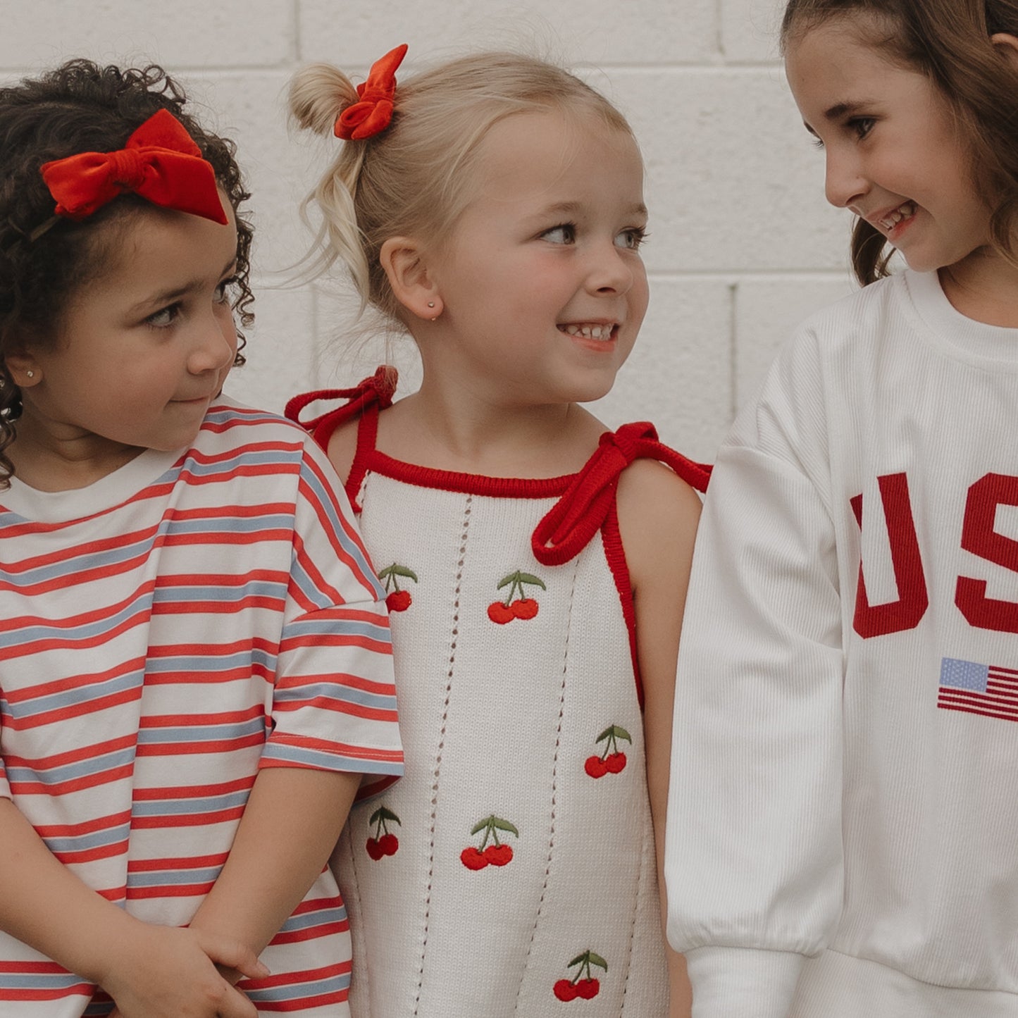 Three smiling girls stand together. One wears a red striped shirt with a headband, another wears the forever french baby Knit Dress | Cherry—100% cotton with cherry prints and tie straps—and the third sports a white “USA” sweatshirt.