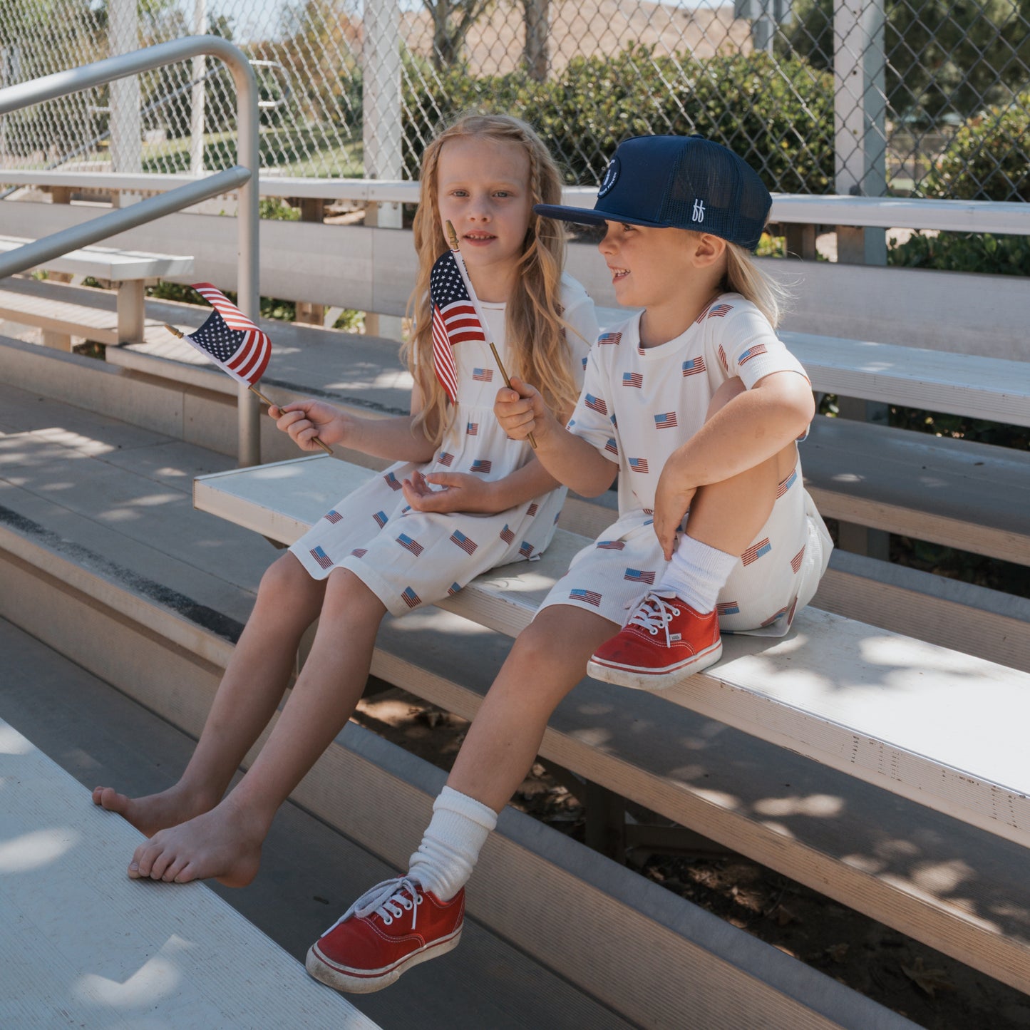 Two children sit on bleachers wearing matching forever french baby Bamboo T-Shirt Dress | Americana Ribbed outfits. One holds a flag, wears a blue cap and red sneakers; the other, also holding a flag, is barefoot.