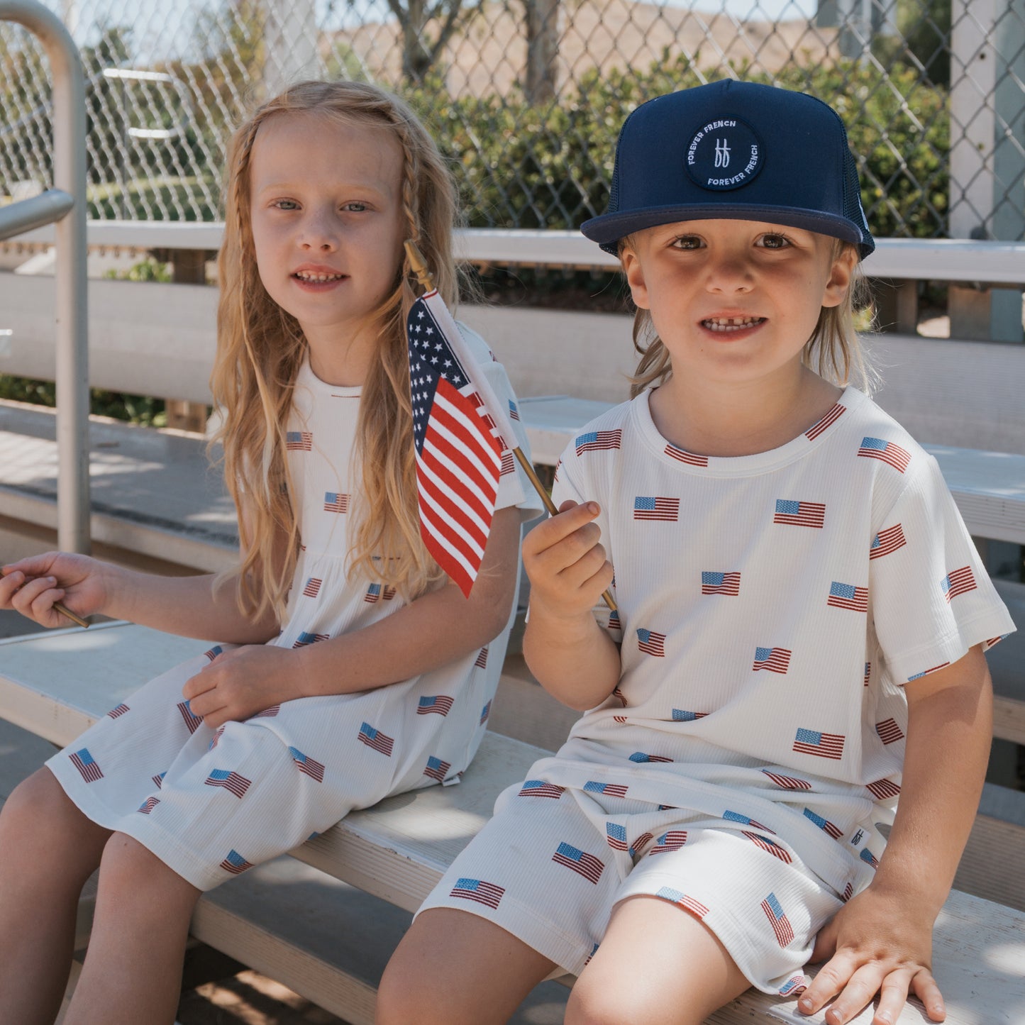 Two young children sit outdoors on bleachers in matching forever french baby Bamboo Short Set | Americana Ribbed outfits featuring small American flag prints. One wears a blue hat and waves a U.S. flag on a sunny, festive day by a chain-link fence.