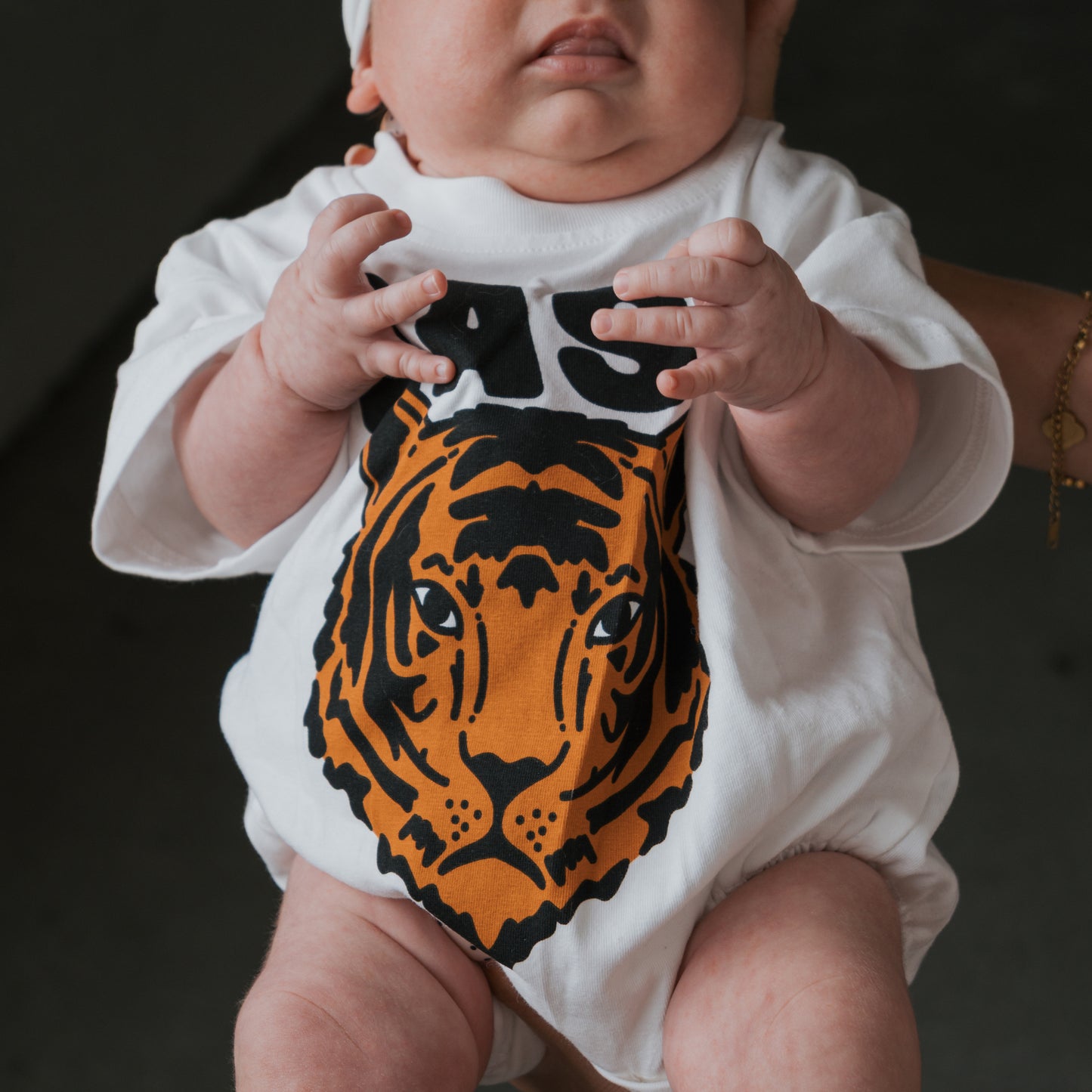 A baby wears the forever french baby "Easy Tiger" short sleeve romper, featuring a white cotton blend and a bold orange and black tiger face design on the front, gently supported by an adult's hand.