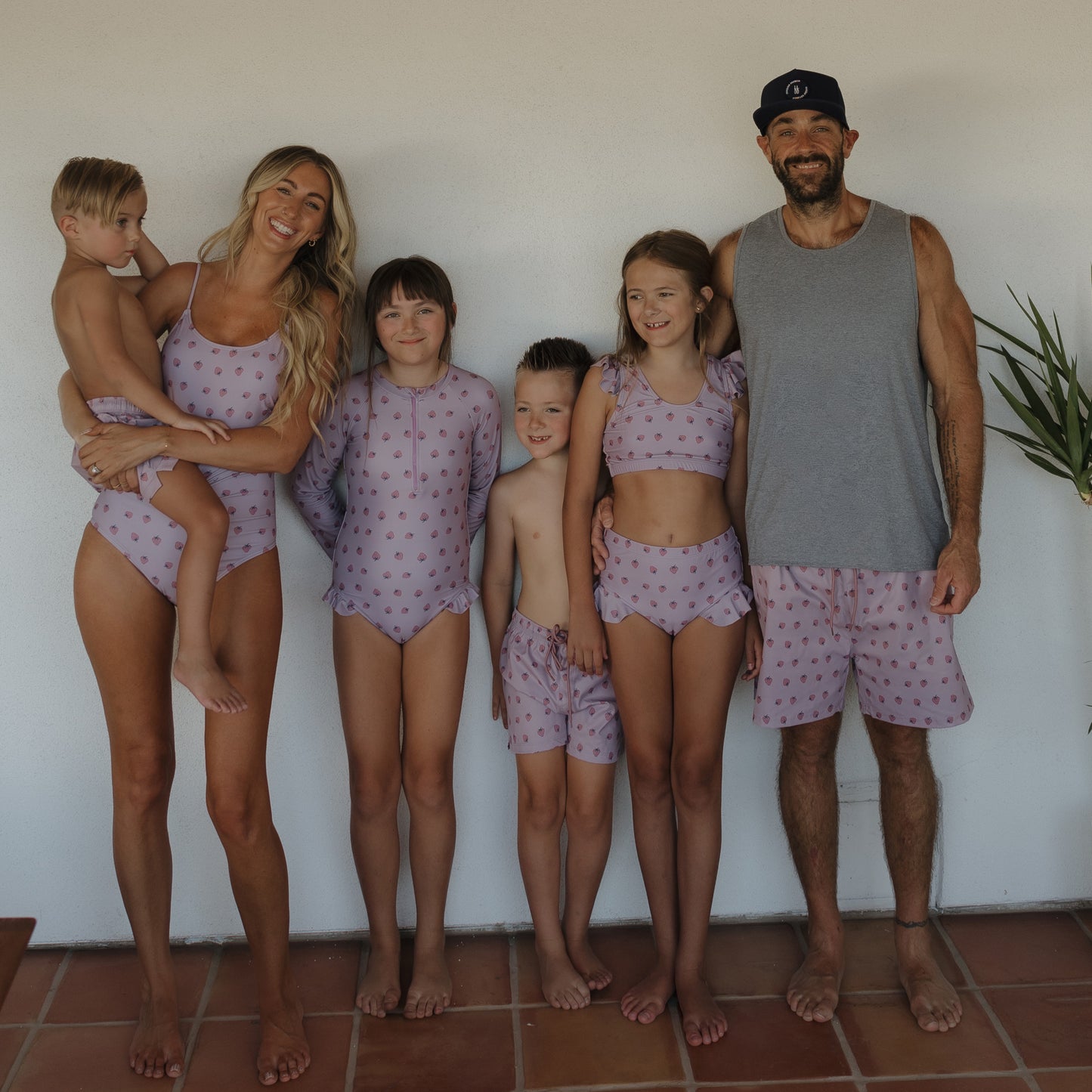 A family of six, including four children, poses barefoot against a white wall in matching forever french baby "Matilda" girl's two-piece swimsuits featuring a light purple color and tiny pink strawberry print.