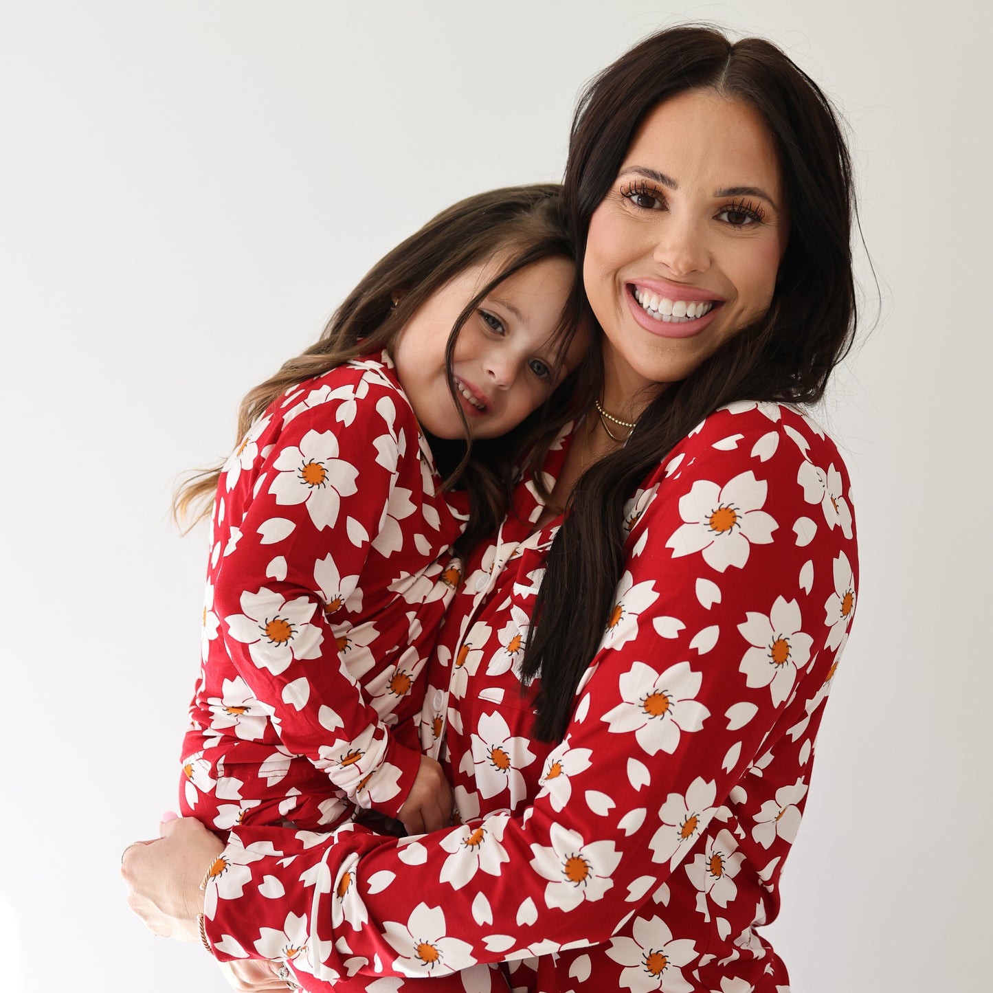 A woman holds a young girl, both smiling and posing in forever french baby’s Girl's Bamboo Button Up Lounge Set in Cherry Blossoms, against a plain white background.
