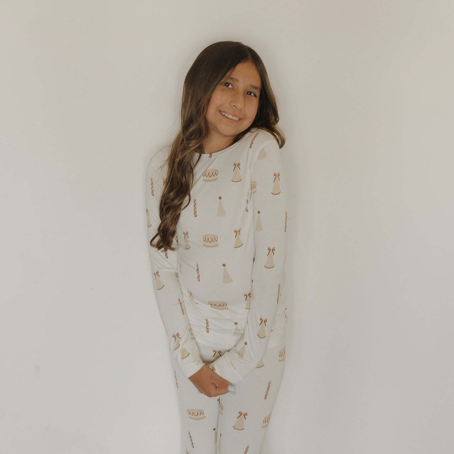 A young girl with long brown hair stands against a plain white wall, smiling gently with her hands clasped in front of her, wearing the forever french baby Child Sleepy Time Set | Birthday Girl—a soft bamboo lounge set featuring matching white pajamas adorned with brown patterns.