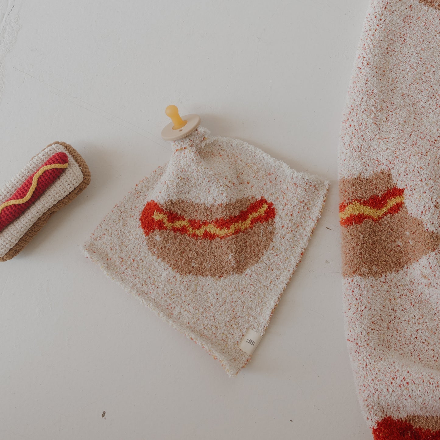 The forever french baby Lovey | Hot Dog is a cozy microfiber feather yarn lovey featuring a cute hot dog design, paired with a matching hot dog-shaped toy and partial blanket in the same pattern, all shown on a white surface.
