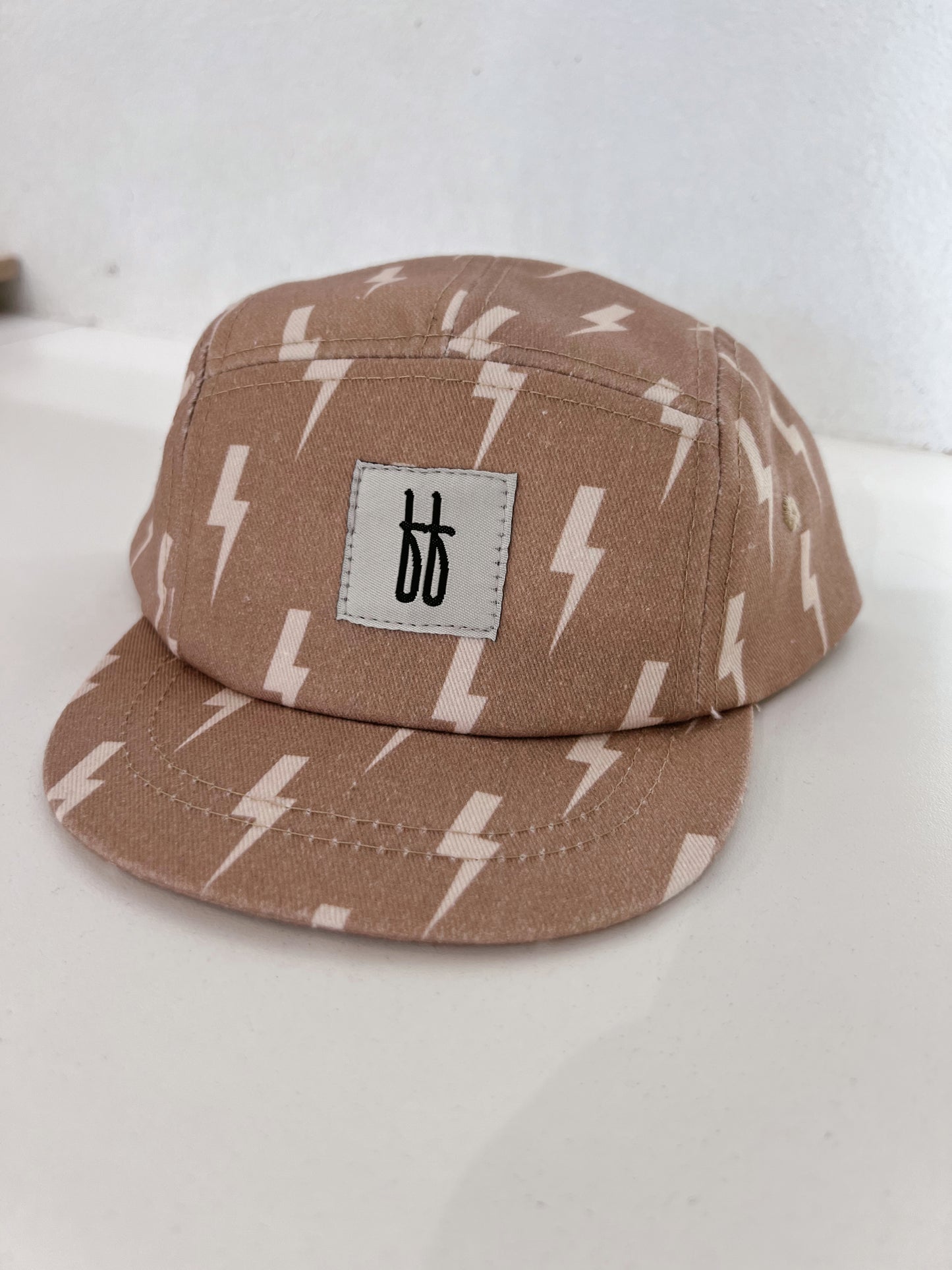 The Snap Back | Tan & Cream Lightning Bolt cap by forever french baby features a pattern of white lightning bolts on a tan 5-panel design. The front panel is adorned with a white patch displaying two overlapping black lowercase "b" letters. This stylish cap rests on a white surface against a plain white background.