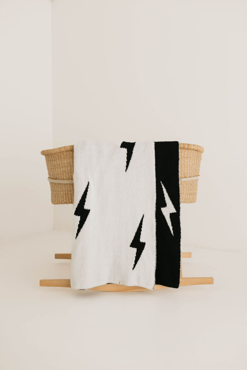 A woven basket sits atop a pale wooden stand against a light-colored backdrop. Draped over the basket is the dreamy soft "Plush Blanket | Black & White Lightning Bolt" by forever french baby, made from 100% Microfiber and Oeko-tex certified, featuring striking black lightning bolt patterns.