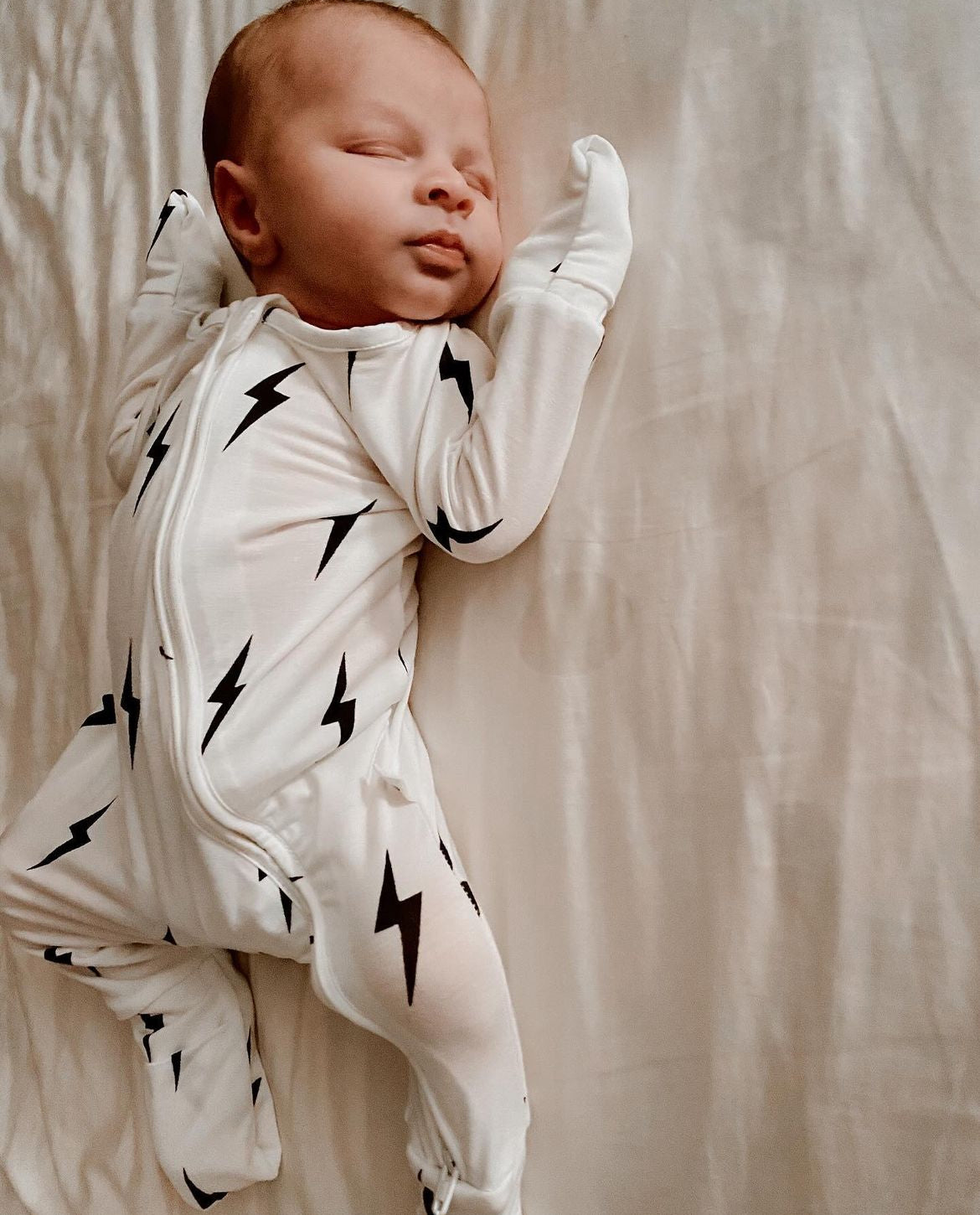 A newborn baby is peacefully sleeping on a bed, dressed in Forever French Baby Bamboo Zip Pajamas in white and black lightning bolt patterns. Their arms are slightly raised above their head, and the light-colored bedding underneath appears incredibly soft.