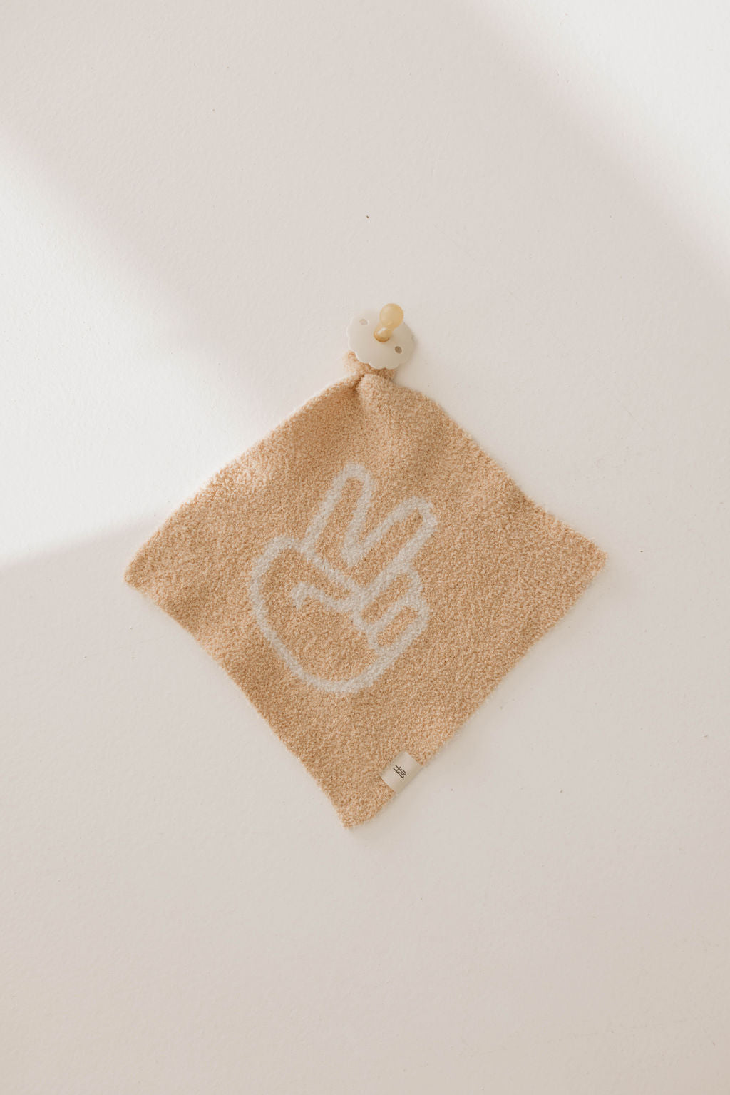 A square beige towel from forever french baby's Lovey | Peace Sign collection hangs on a white wall. This towel, reminiscent of a peaceful warrior's banner, features a white peace sign design, has a small gray tag at the bottom, and is attached to a wooden button.