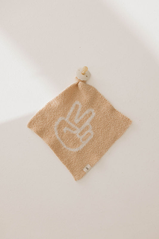 A square beige towel from forever french baby's Lovey | Peace Sign collection hangs on a white wall. This towel, reminiscent of a peaceful warrior's banner, features a white peace sign design, has a small gray tag at the bottom, and is attached to a wooden button.