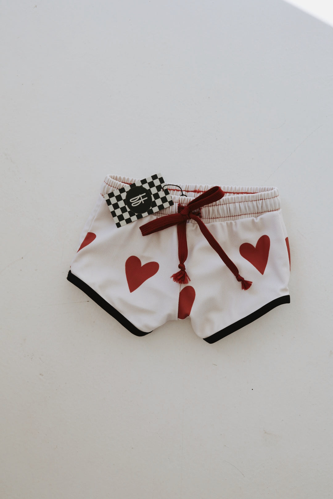 The "Boys Board Short | King of Hearts" from forever french baby, featuring white fabric adorned with red heart patterns, red drawstrings, and black trim, is laid flat on a white background. A black tag with a white symbol and a black-and-white checkered pattern is attached to the waistband. These stylish shorts also offer UPF 50+ protection for those sunny days.
