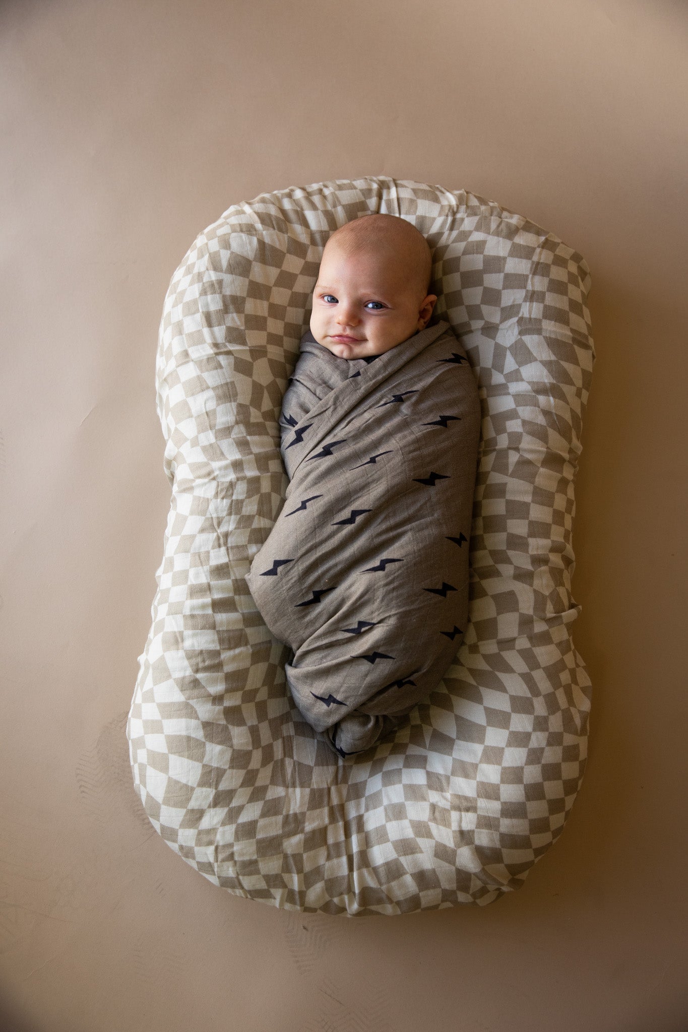 A baby wrapped in a Forever French Baby muslin swaddle in the Wavy Checker pattern is lying in a checkered baby lounger. The baby is looking up with a content expression, set against a plain, neutral-colored background.