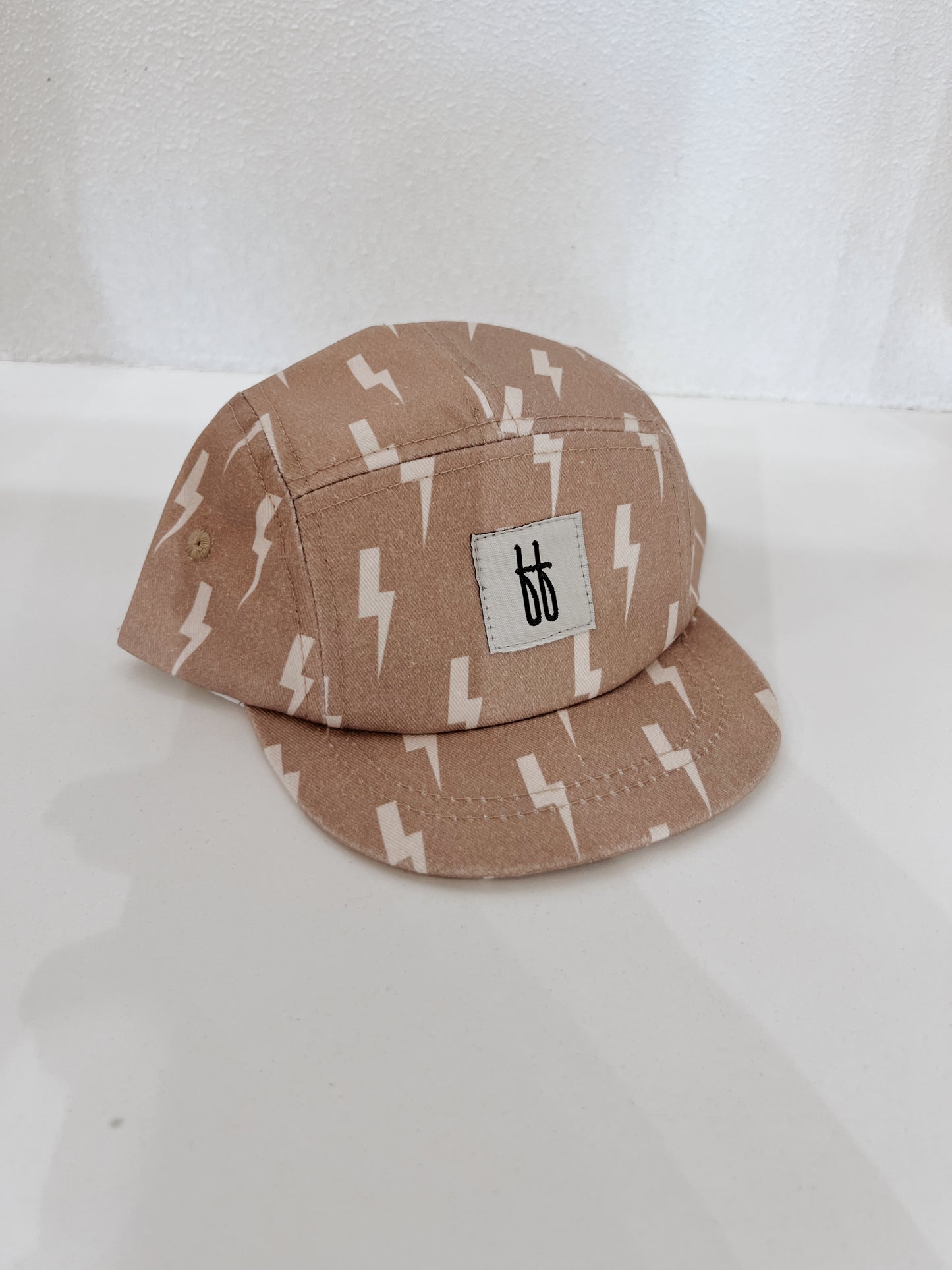 The Snap Back | Tan & Cream Lightning Bolt cap from forever french baby showcases a beige design with a repeating lightning bolt print and a small rectangular patch featuring black "bb" initials on a white background. The cap is photographed on a white surface against a white textured backdrop.