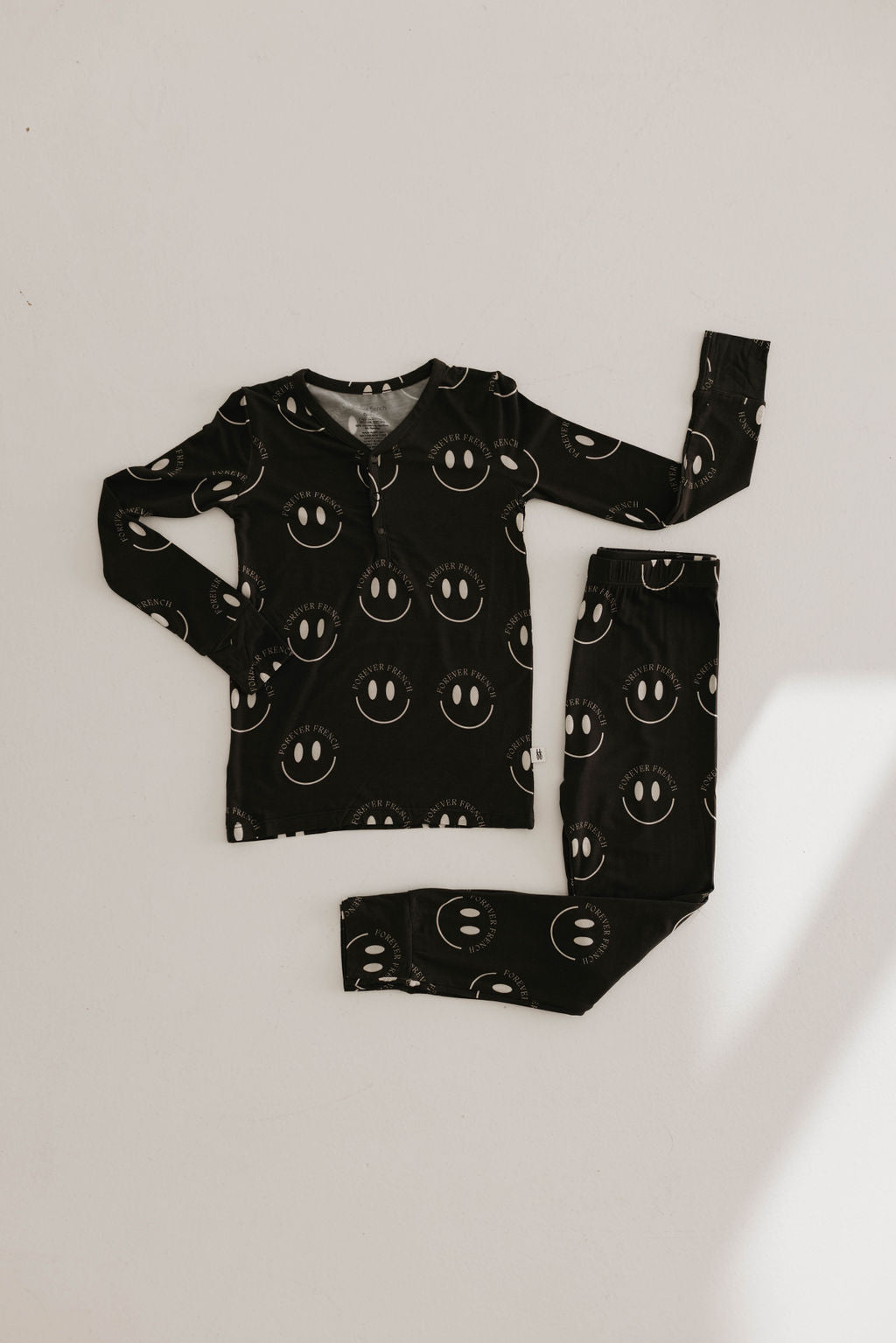 A set of Bamboo Two Piece Pajamas in Charcoal & White ff Smile by forever french baby is laid flat on a light background. The sleepwear, featuring a long-sleeve top and pants both with white smiley face patterns, is designed to be hypo-allergenic. The clothing items are neatly arranged, with the top's sleeves and pants slightly bent.