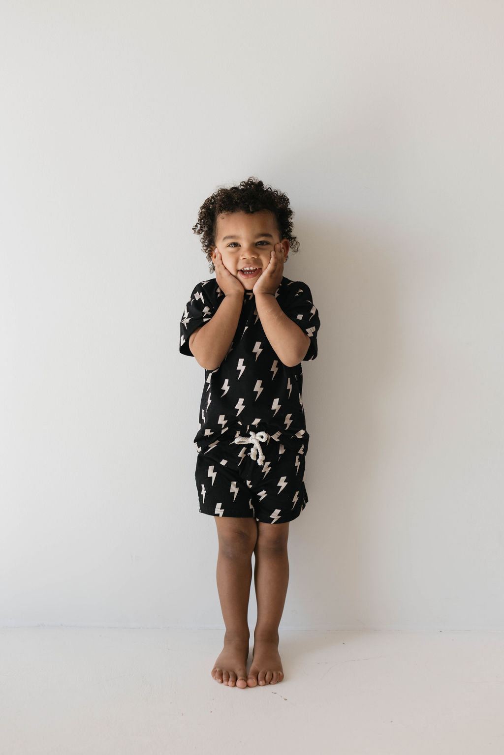 A young child with curly hair stands barefoot against a plain backdrop, wearing the Short Set | Black Lightning from forever french baby. The child, aged 0-9 years, is smiling and holding their cheeks with both hands in an expressive, joyful gesture.