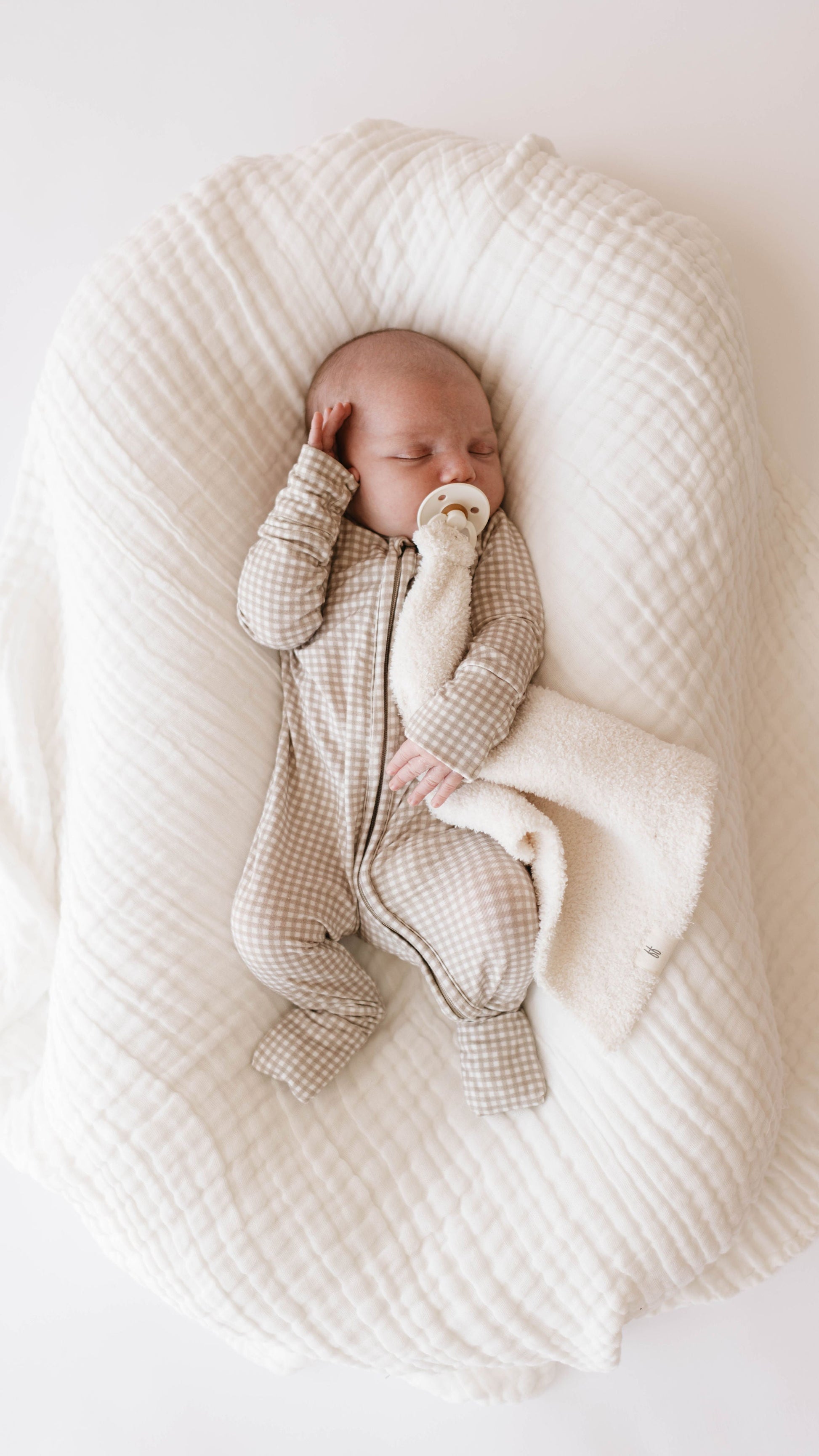 An infant is lying on a soft, white Quilt | Cloud blanket from Forever French Baby. The baby is dressed in a gray and white checkered onesie, holding a pacifier and a beige plush blanket. With one hand up by its head, the baby appears to be peacefully sleeping. For best care, wash with like colors.