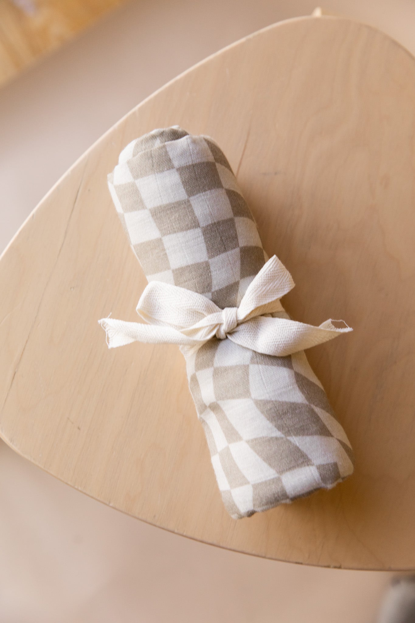 A fabric bundle, wrapped in a forever french baby Muslin Swaddle | Wavy Checker, secured with a white ribbon, sits on a light wooden tabletop. The background is softly blurred.