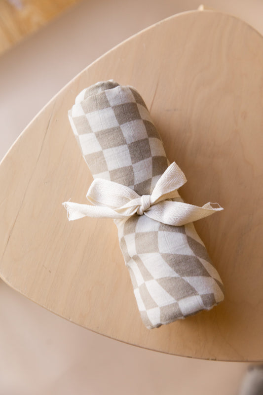 A fabric bundle, wrapped in a forever french baby Muslin Swaddle | Wavy Checker, secured with a white ribbon, sits on a light wooden tabletop. The background is softly blurred.