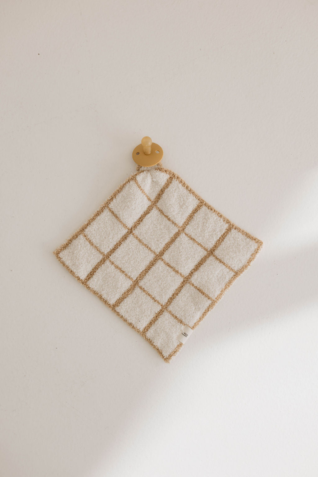A beige and white checkered baby washcloth, named Lovey | Golden Grid from forever french baby, lies flat on a white surface with a beige baby pacifier attached at one corner. Made from Forever French Baby's microfiber feather yarn, the washcloth has a soft, textured appearance and features a small tag on one edge.