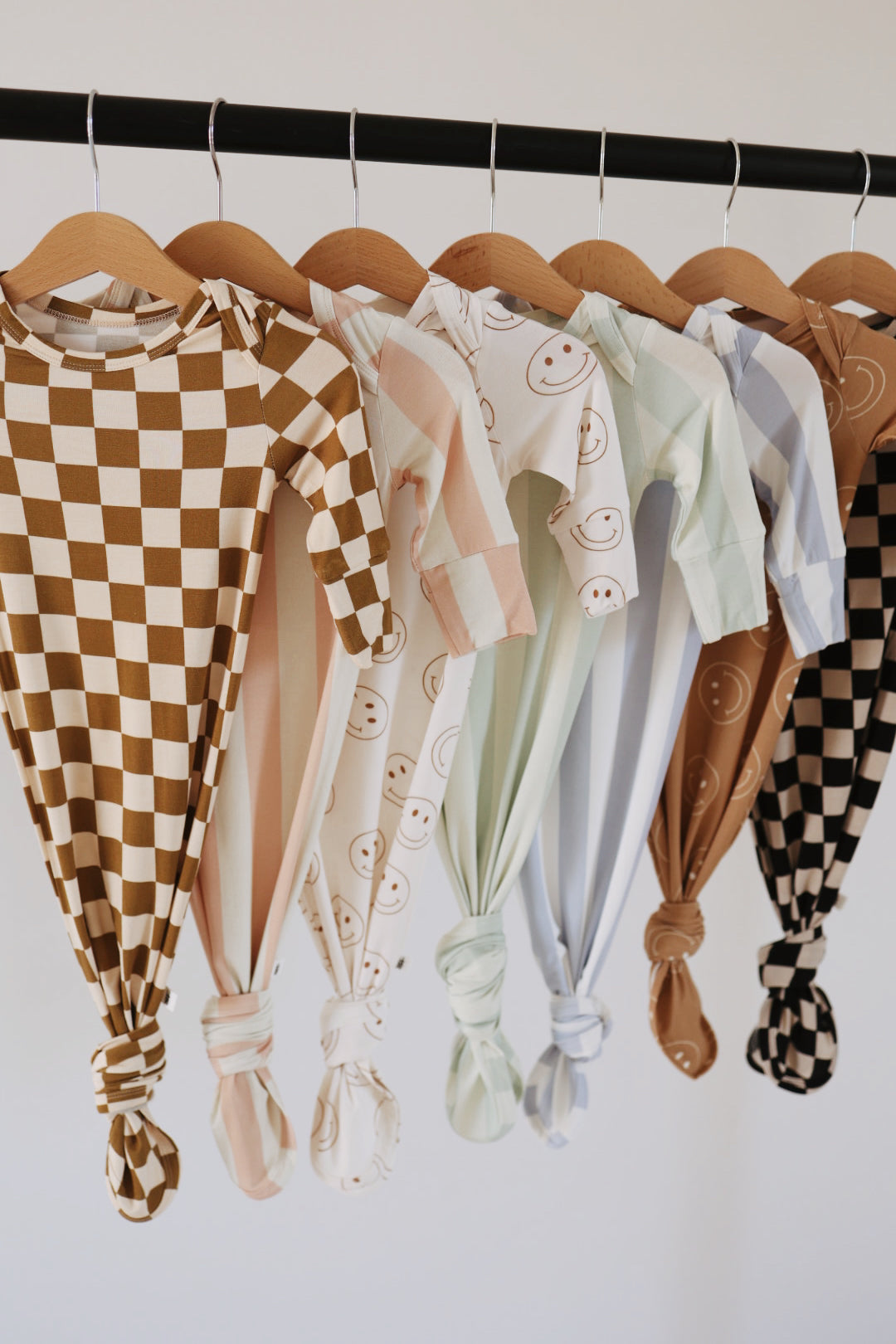 A row of Forever French Baby knotted gowns, made from hypo-allergenic bamboo fabric, hanging from wooden hangers. The gowns feature an array of pastel colors and patterns, including brown checkerboard, peach and white stripes, smiley faces, mint green, light blue, beige, and black and brown checkerboard.