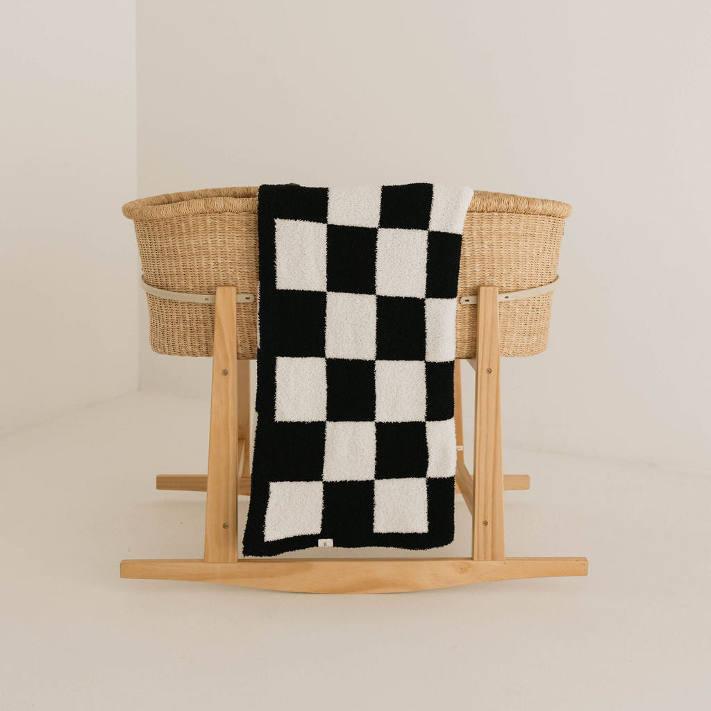 A woven bassinet stands in a minimalist room. Draped over one side is a Plush Blanket in a black and white checkerboard pattern by Forever French Baby, certified to the OEKO-TEX Standard 100. The overall setting features neutral colors and simple decor, highlighting the natural texture of the woven bassinet and the bold pattern of the soft blanket.